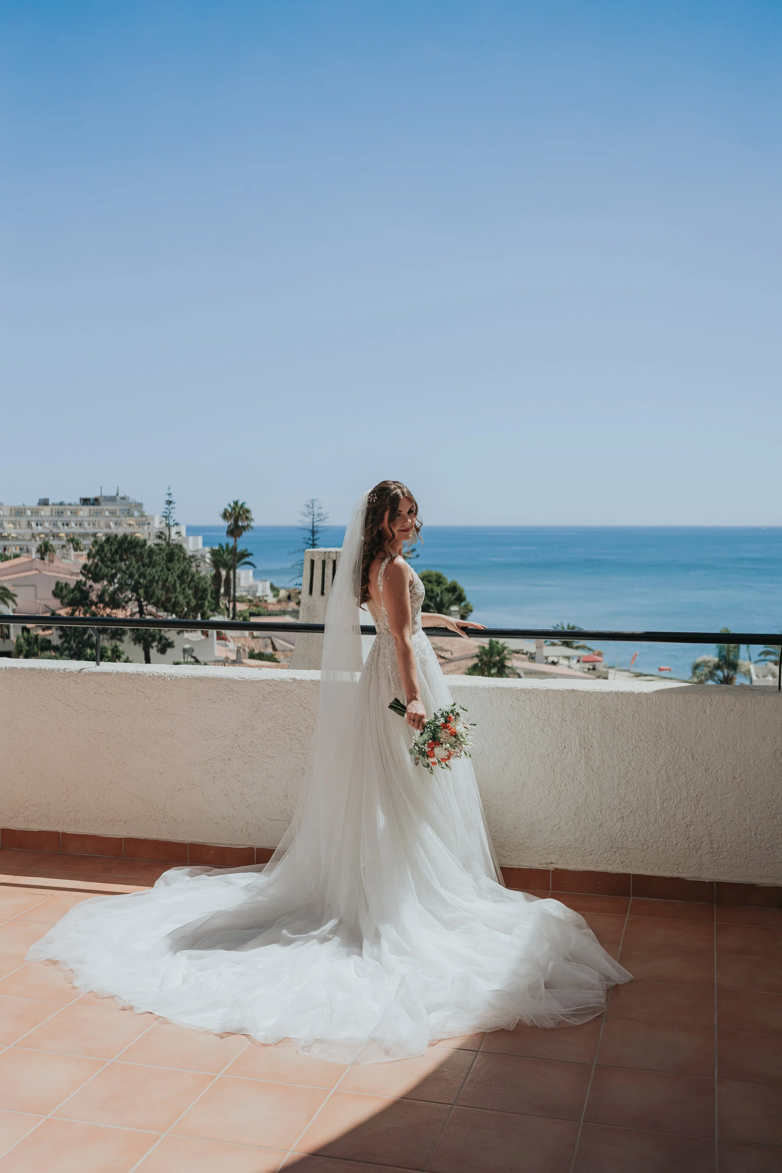 A bride in a white wedding gown holding a bouquet of flowers, standing on a balcony with the ocean and clear blue sky in the background.