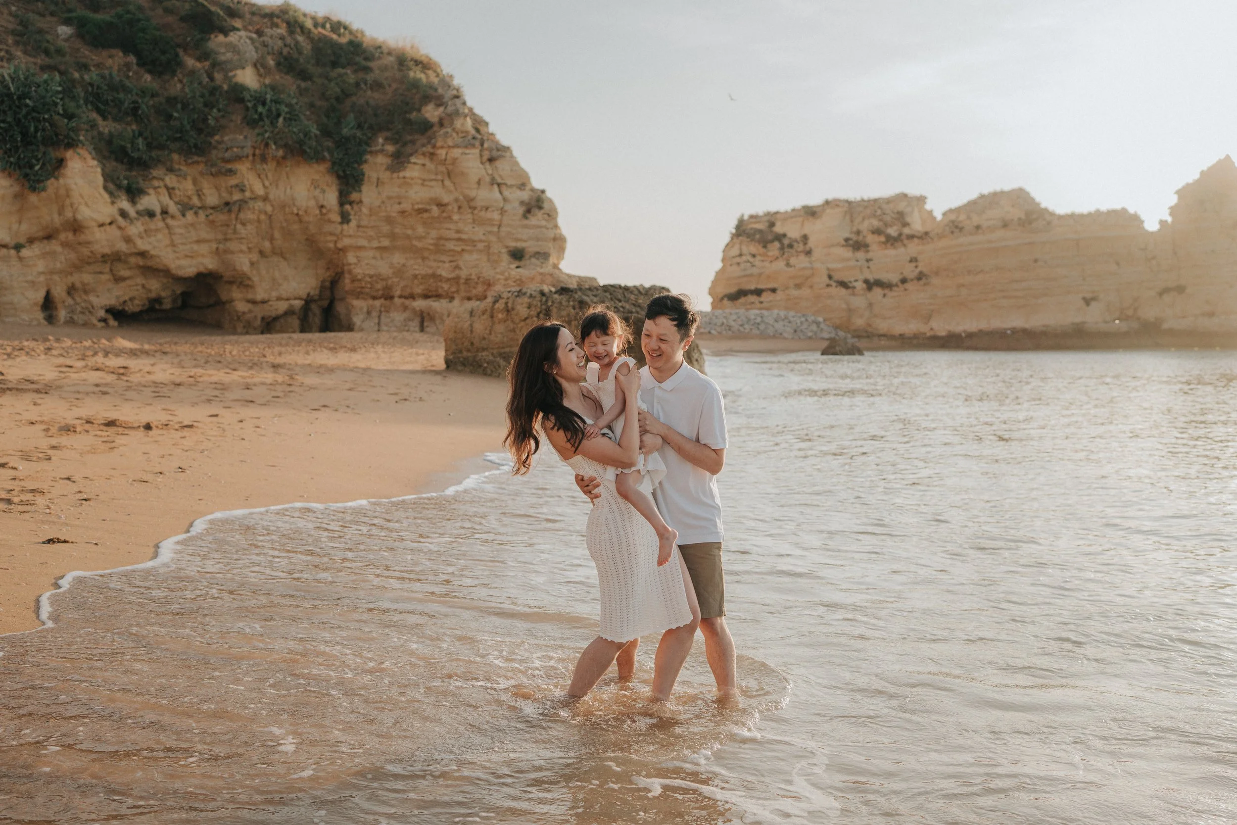 A family of three enjoying a walk on the beach in the late afternoon sunlight. The mother, father, and young girl are smiling and playing close to the shoreline with rugged cliffs in the background.