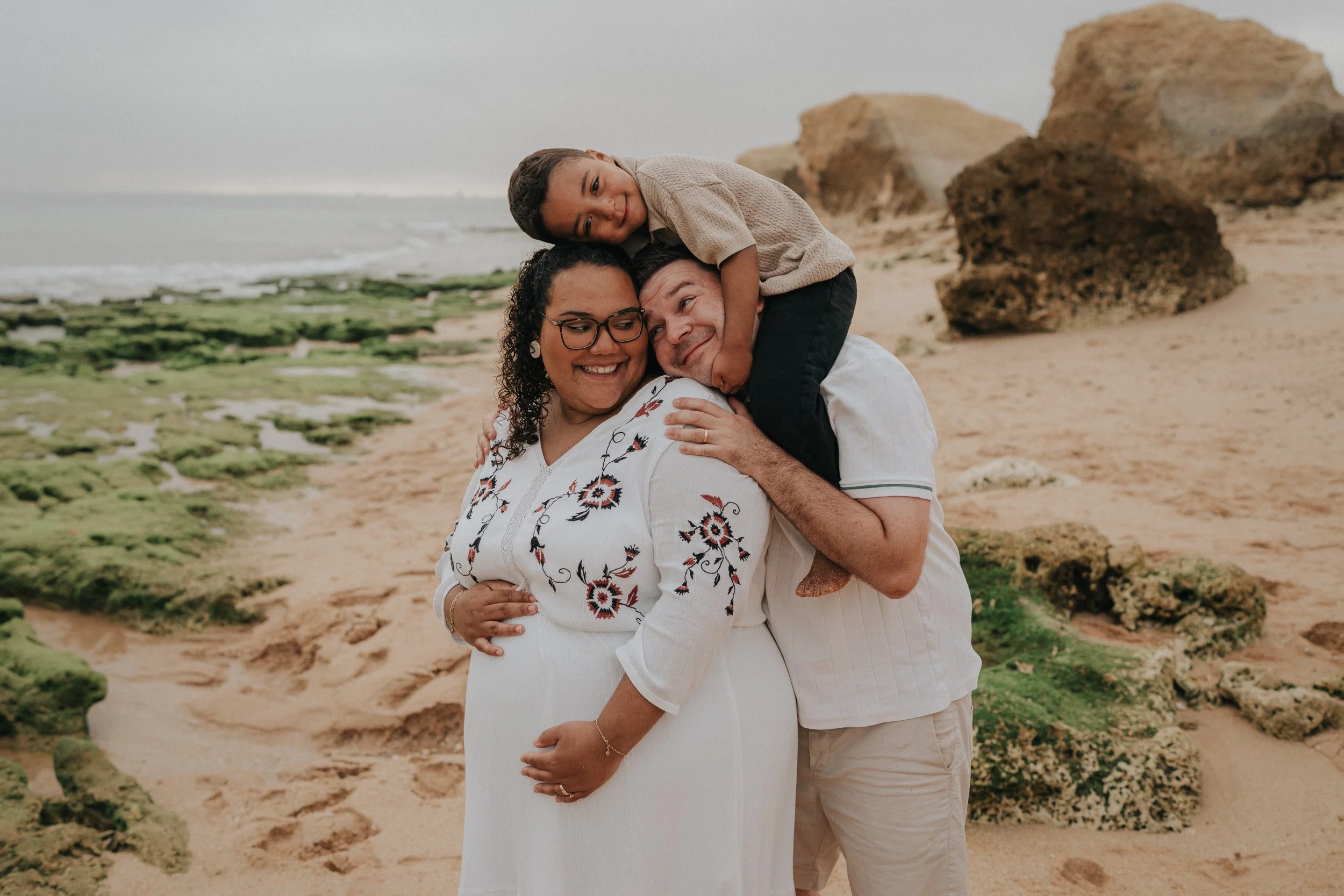 A happy family of three at the beach, with rocky formations and ocean waves in the background. The woman is smiling, the man is hugging her, and the girl is resting her head on the man's head.