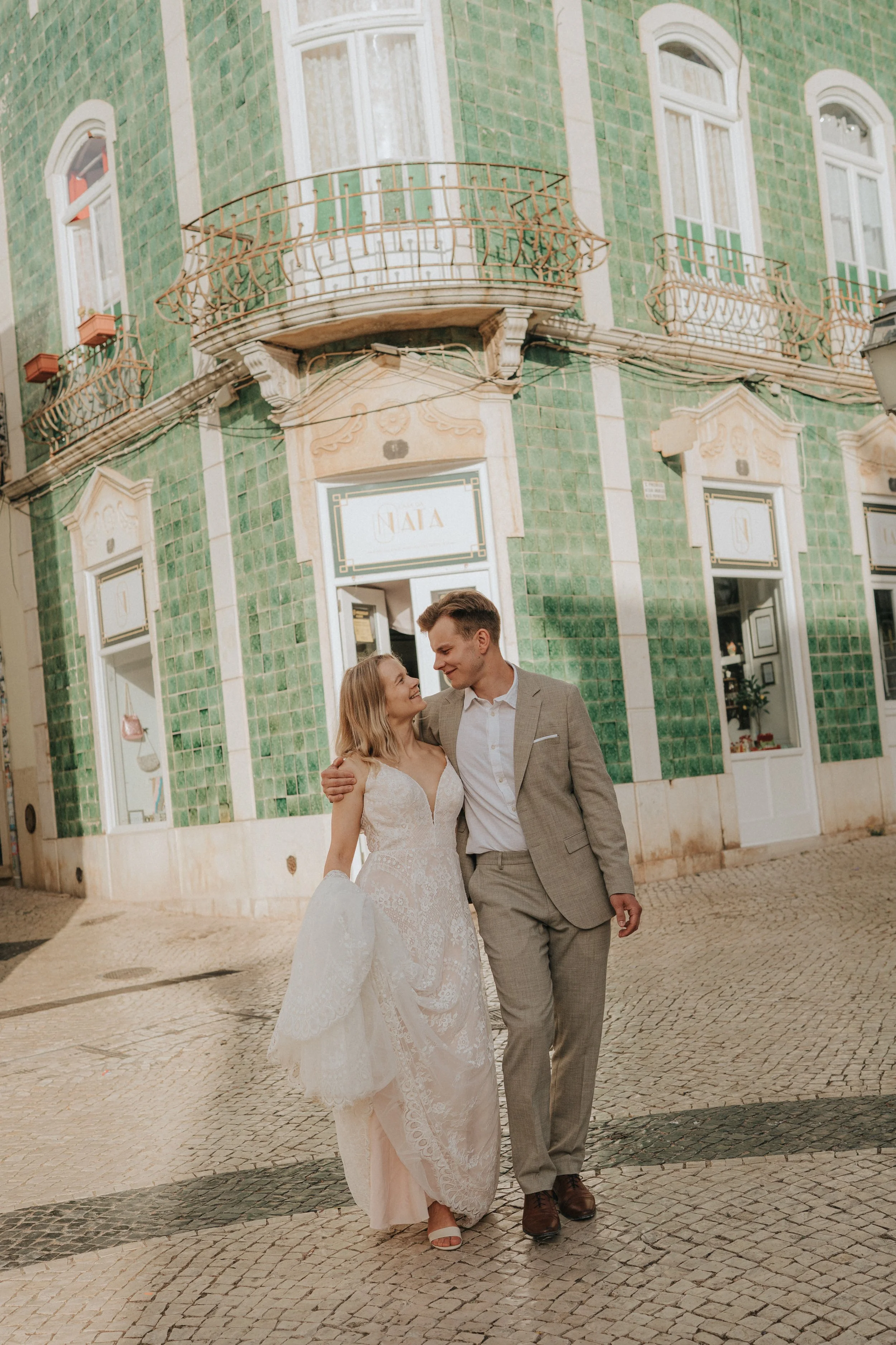 A bride and groom walk together outside a green-tiled building with white decorative details. The bride wears a white lace wedding dress and carries a bouquet, while the groom wears a light gray suit.