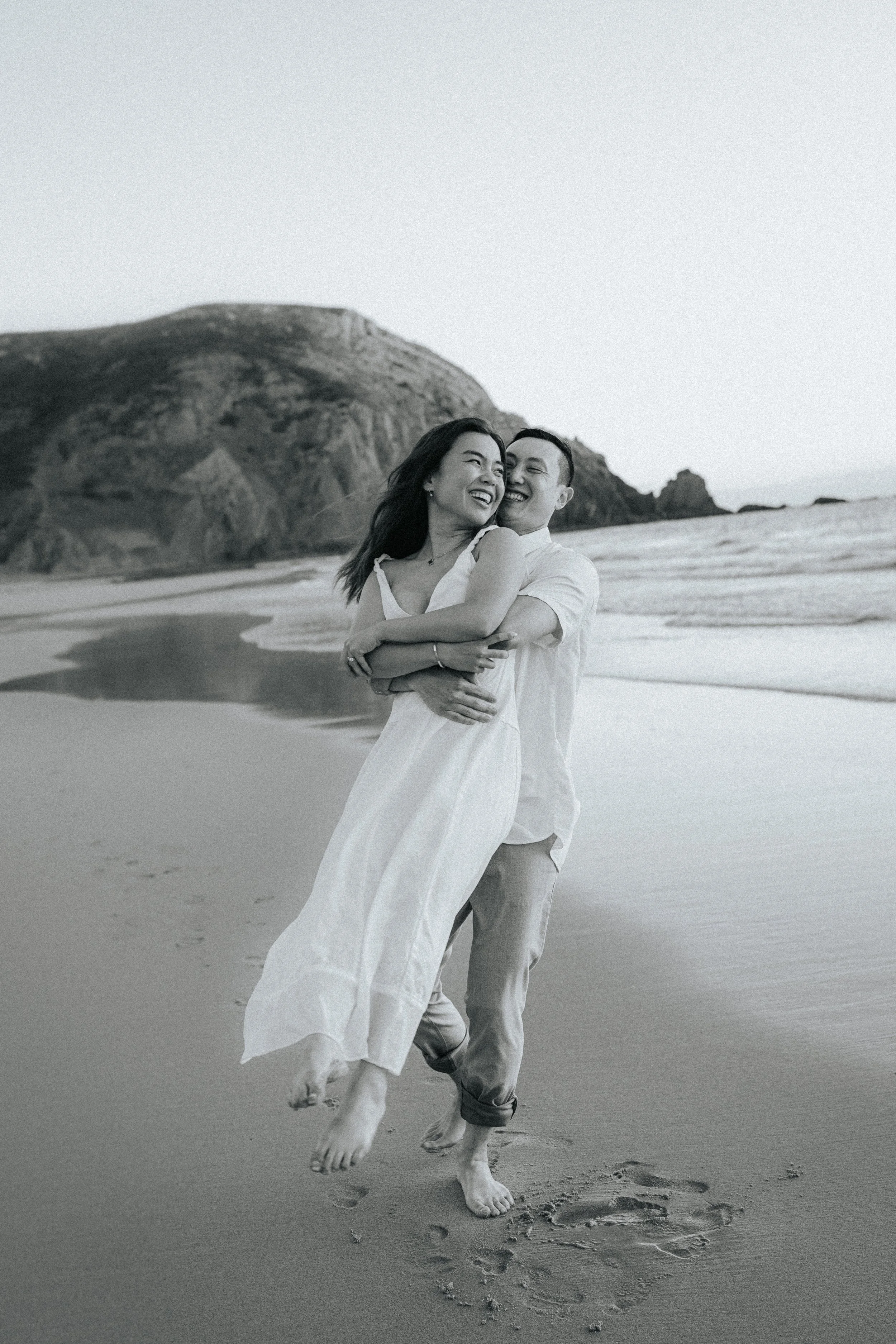 A happy couple, a man and a woman, are running on the beach, holding each other and smiling, with footprints in the sand and a mountainous landscape in the background.