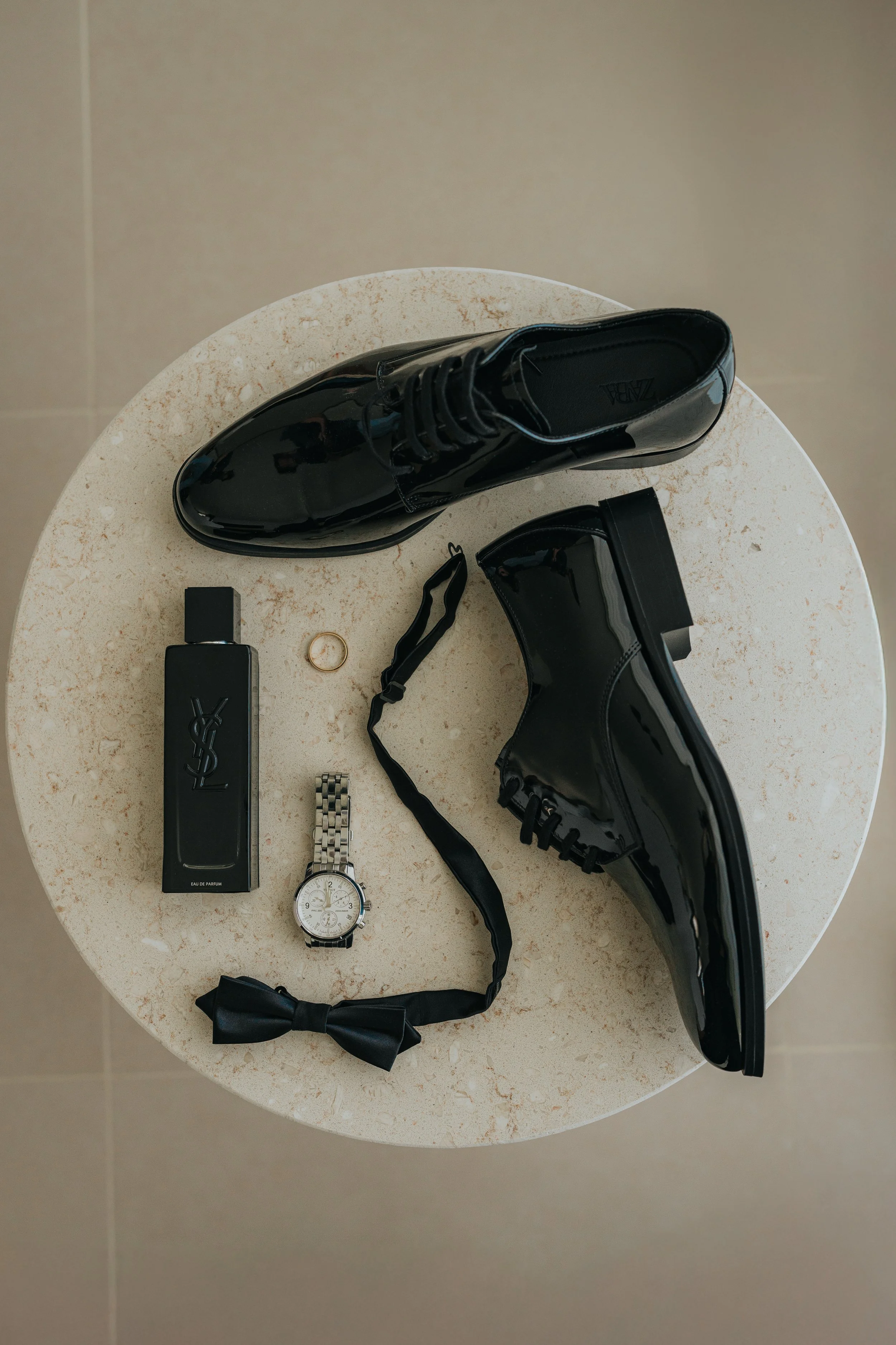 A round table with various items on it, including a black high-heeled shoe, a black perfume bottle, a silver wristwatch, a black bow tie, a gold ring, a black strap, and a pair of black ankle boots.