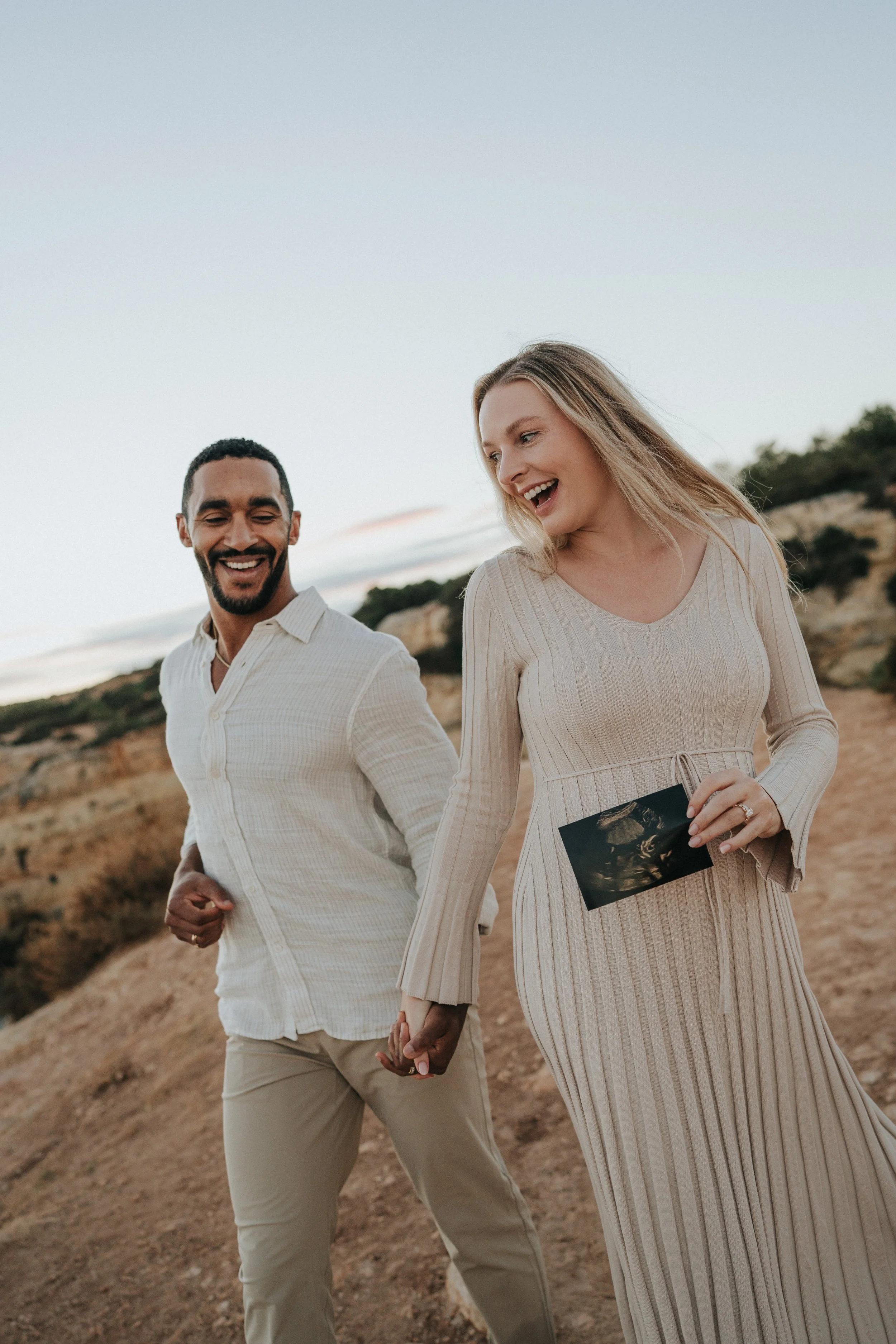 A couple walking hand in hand outdoors, woman holding an ultrasound picture, smiling and looking at each other, landscape background with hills and sky.
