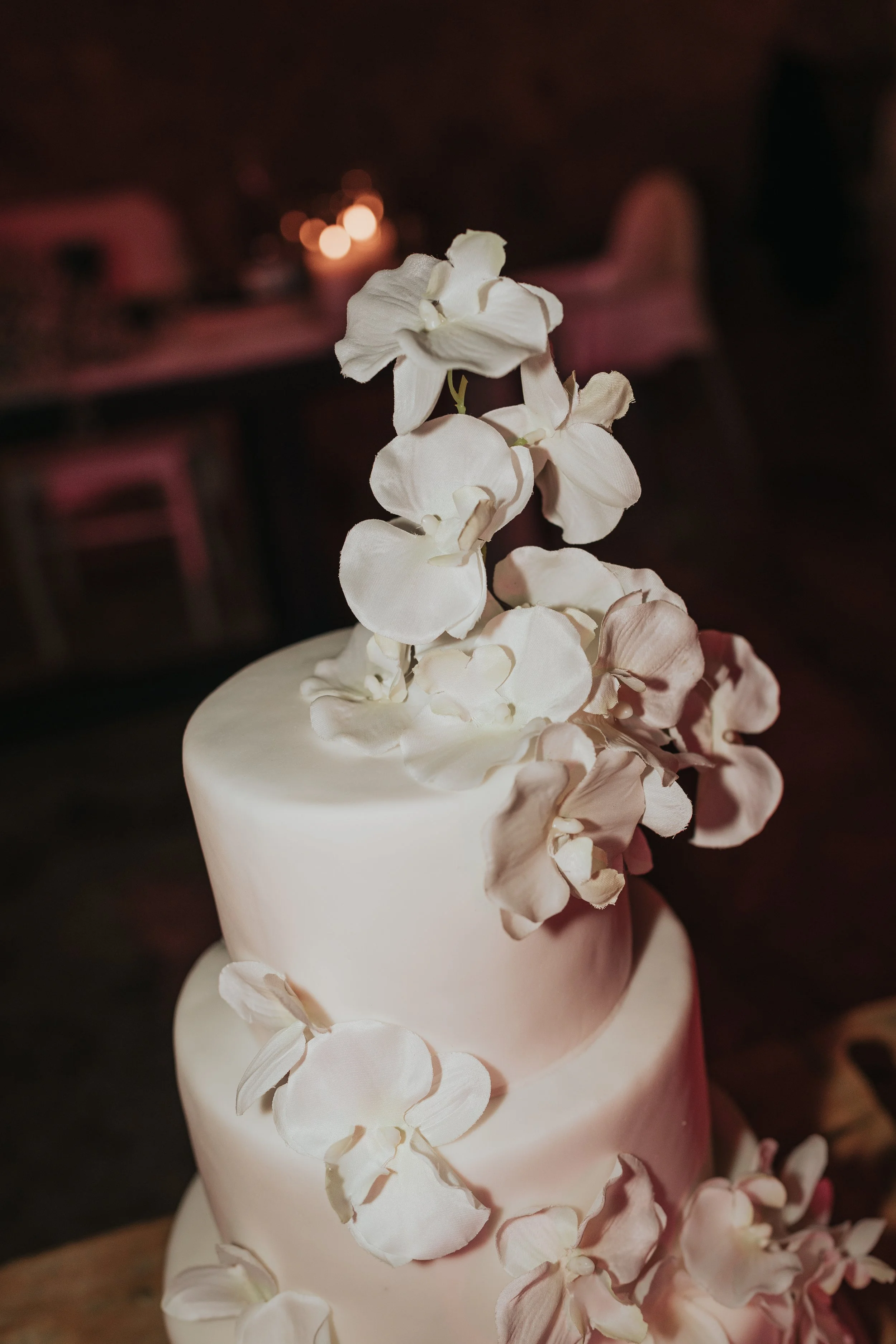 A white ceramic vase holding a white floral arrangement with large, soft petals, set against a blurred dark background with warm lights.