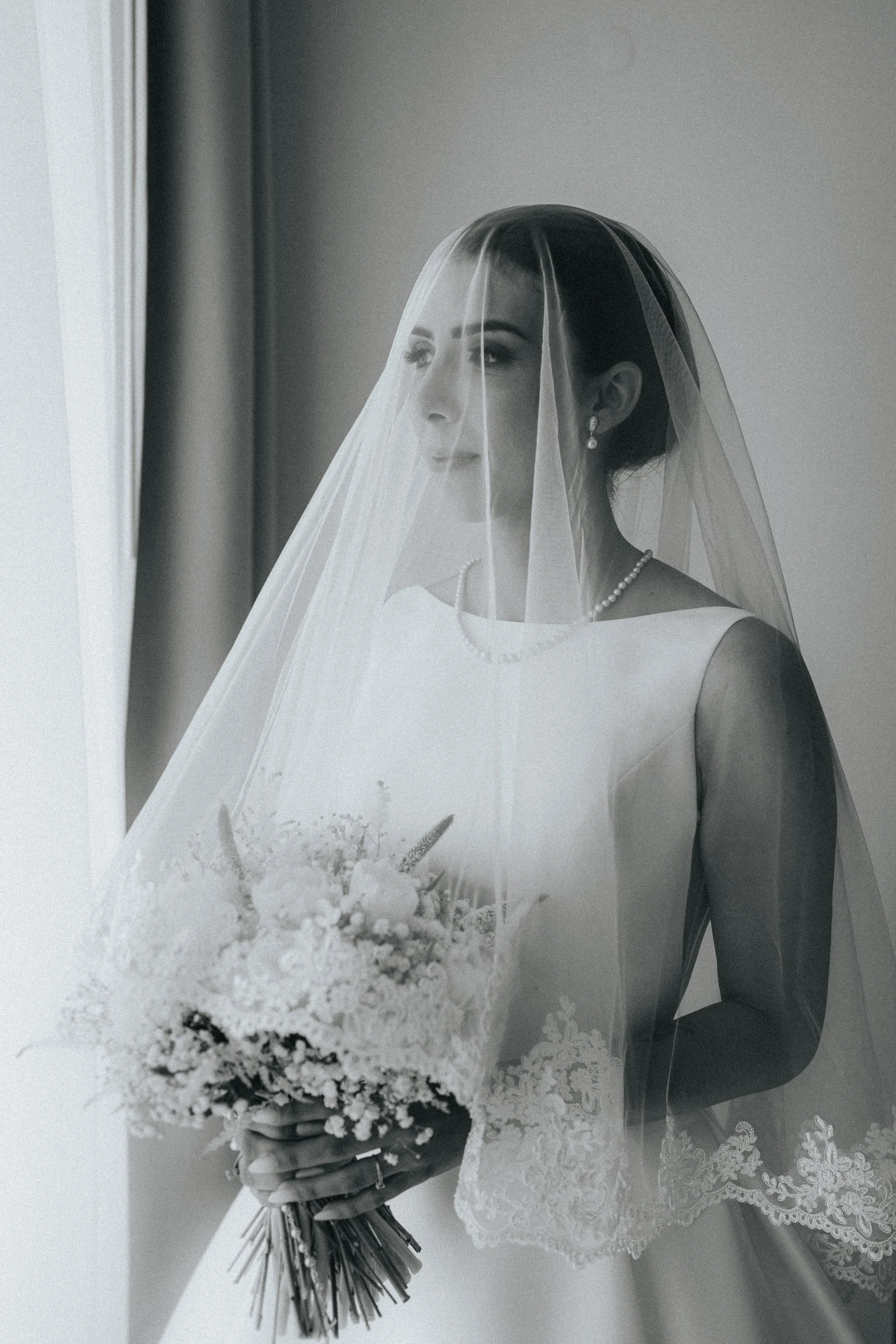 A bride wearing a wedding dress, pearl necklace, and earrings, with her face partially covered by a veil, holding a bouquet of flowers.