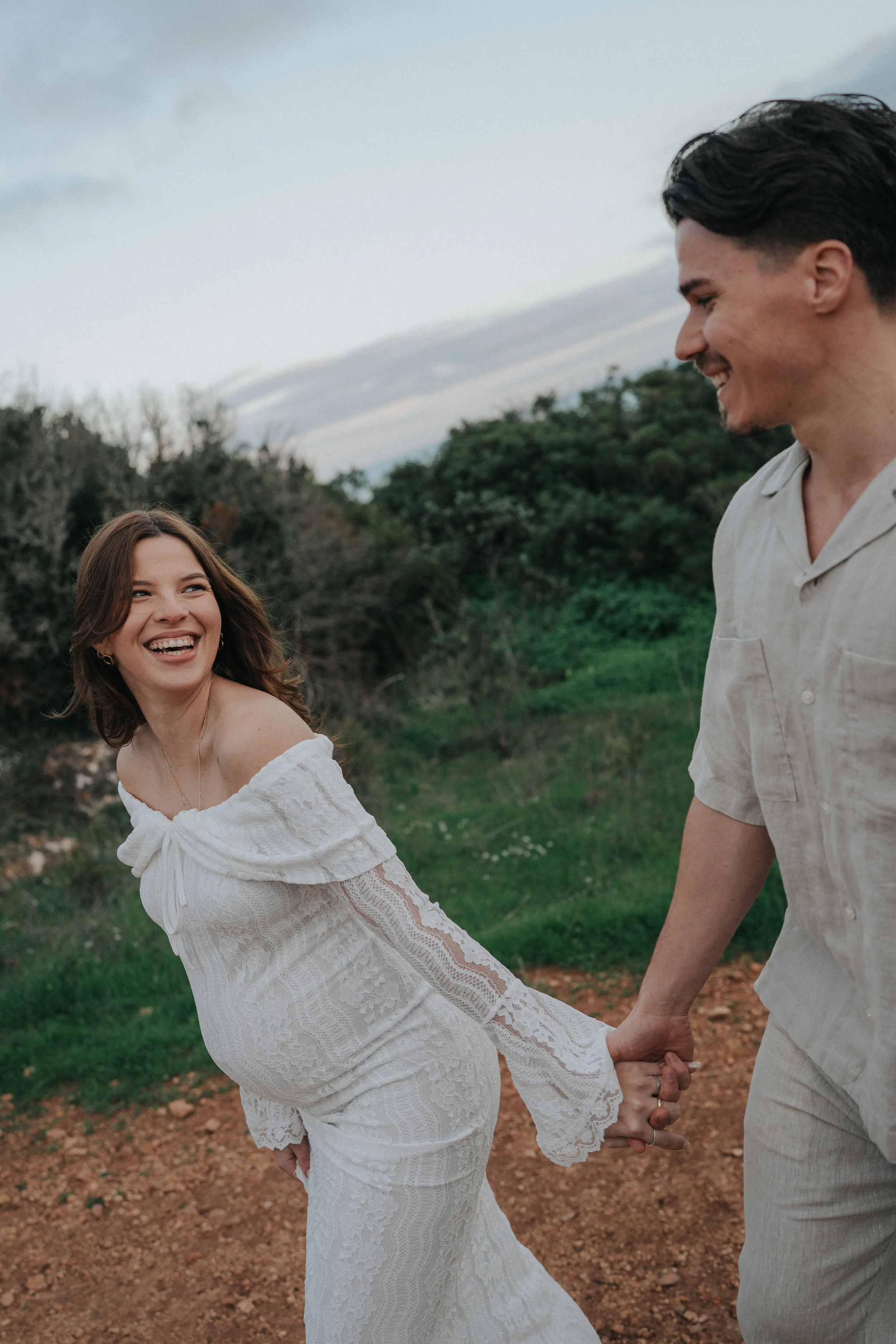 A man and a woman are smiling and holding hands outdoors. The woman is wearing a white lace dress, and the man is wearing a light-colored shirt and pants. They are standing on a dirt path with greenery and trees in the background, and it appears to b