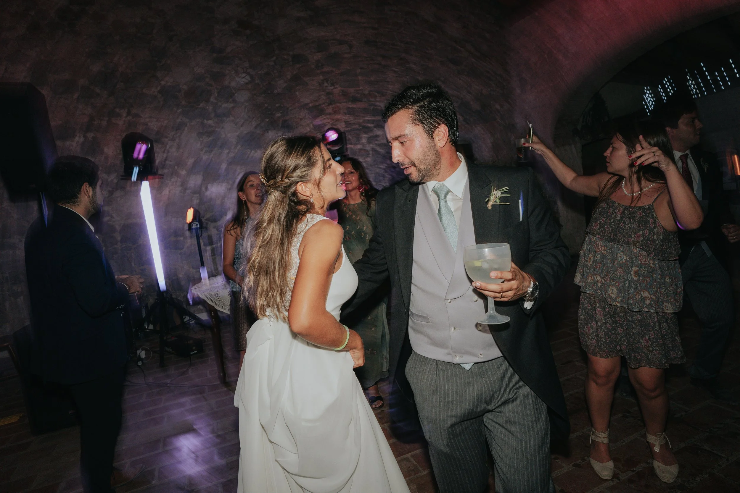 A bride and groom dancing at their wedding reception surrounded by friends in a dimly lit venue with brick walls and purple accent lighting.
