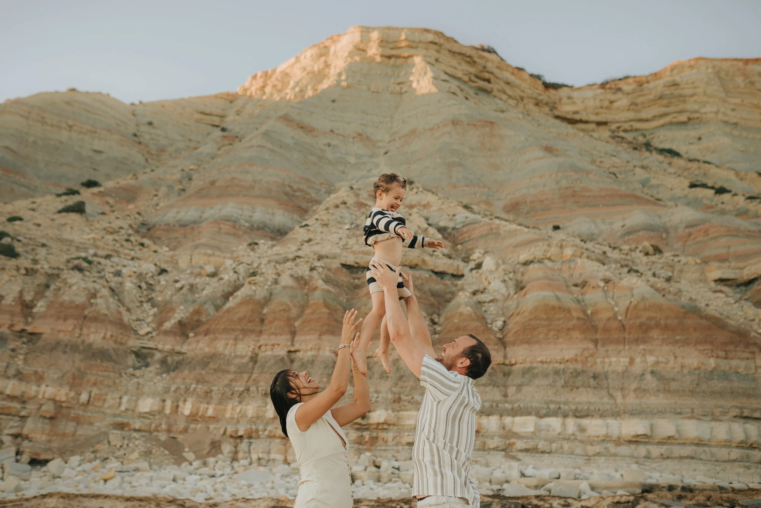 A family of three at the beach with a layered mountain in the background. The father is lifting a smiling child in the air, and the mother is reaching up to hold the child's hand.