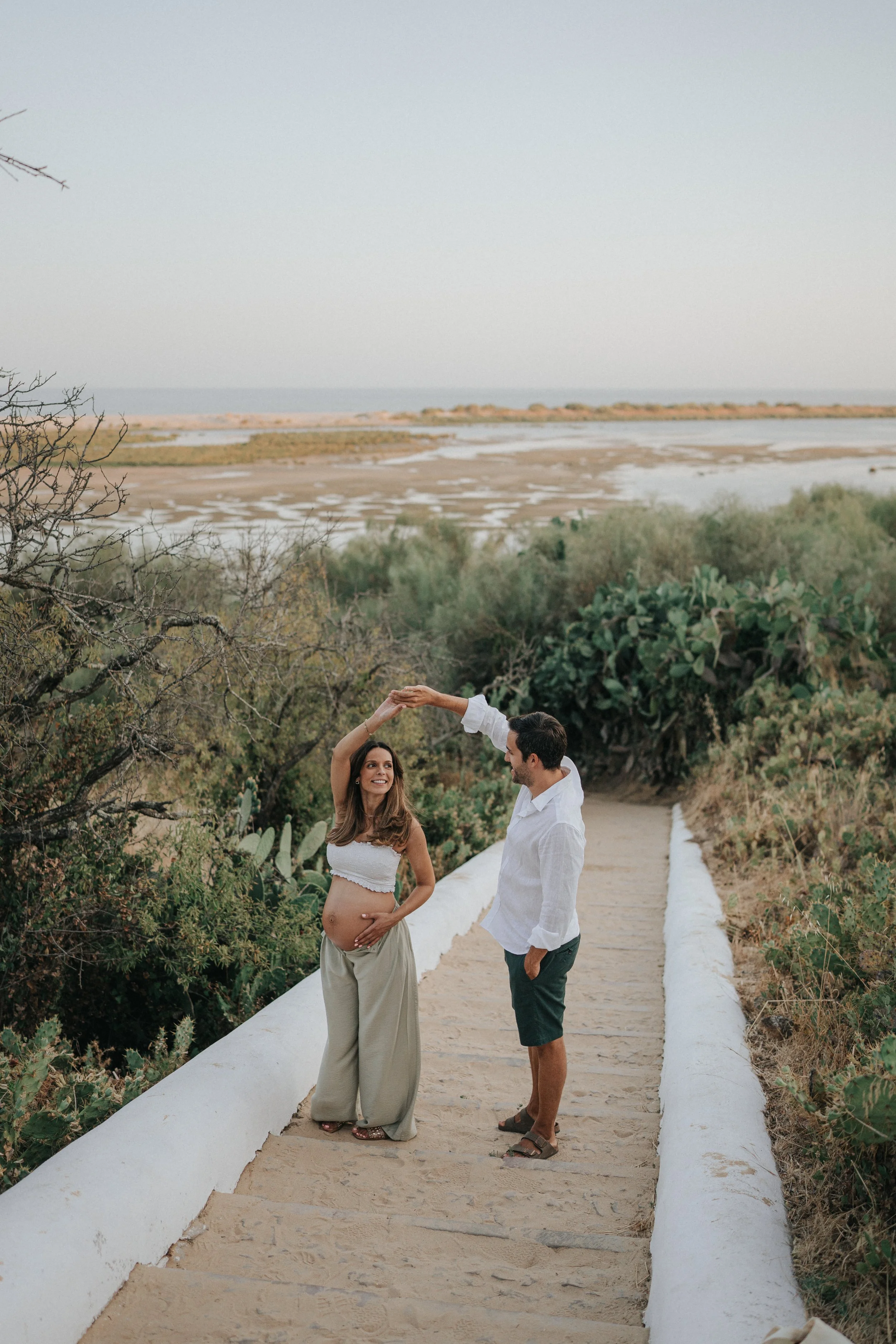 A pregnant woman and a man are standing on a sandy pathway in a natural, outdoor setting with green bushes and trees. The woman is wearing a white crop top and beige pants, holding her belly, and the man is dressed in a white shirt and dark shorts. T