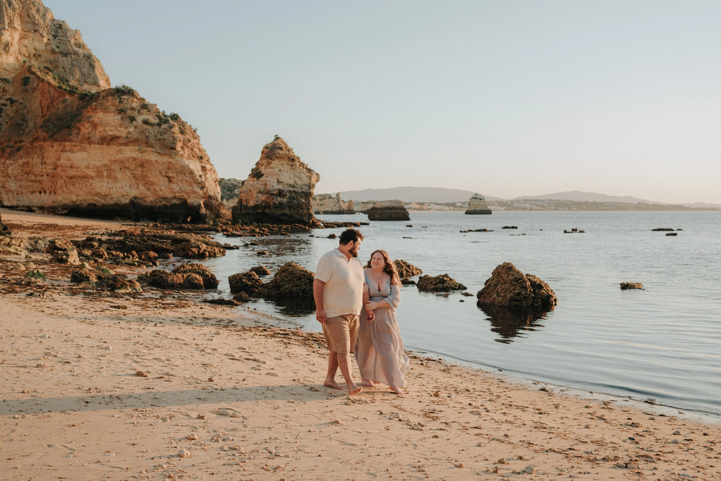 A couple walking along a sandy beach with large rock formations and cliffs in the background during sunset.