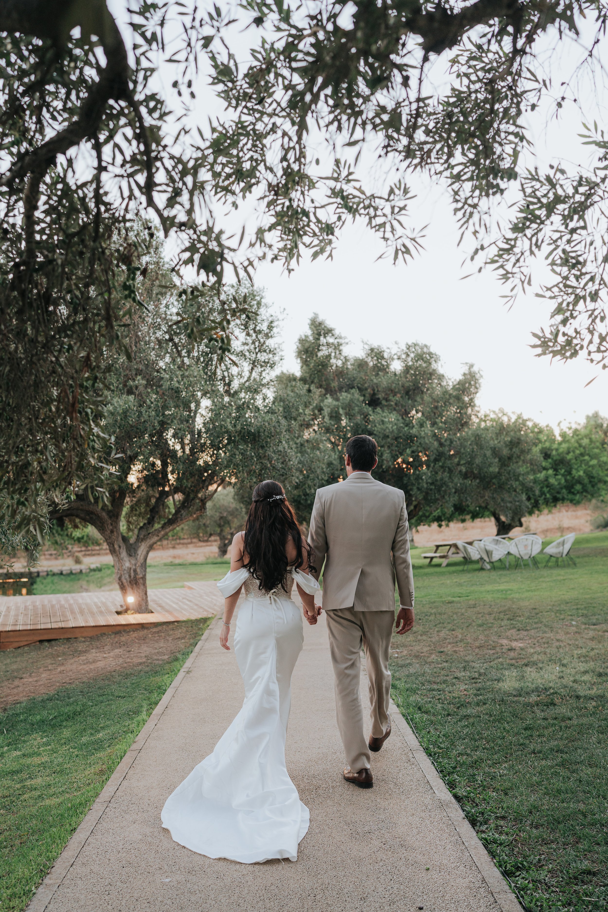 A couple, holding hands, walking away on a paved path through a park with green grass and trees, during sunset. The woman is wearing a white wedding dress and the man is in a beige suit.