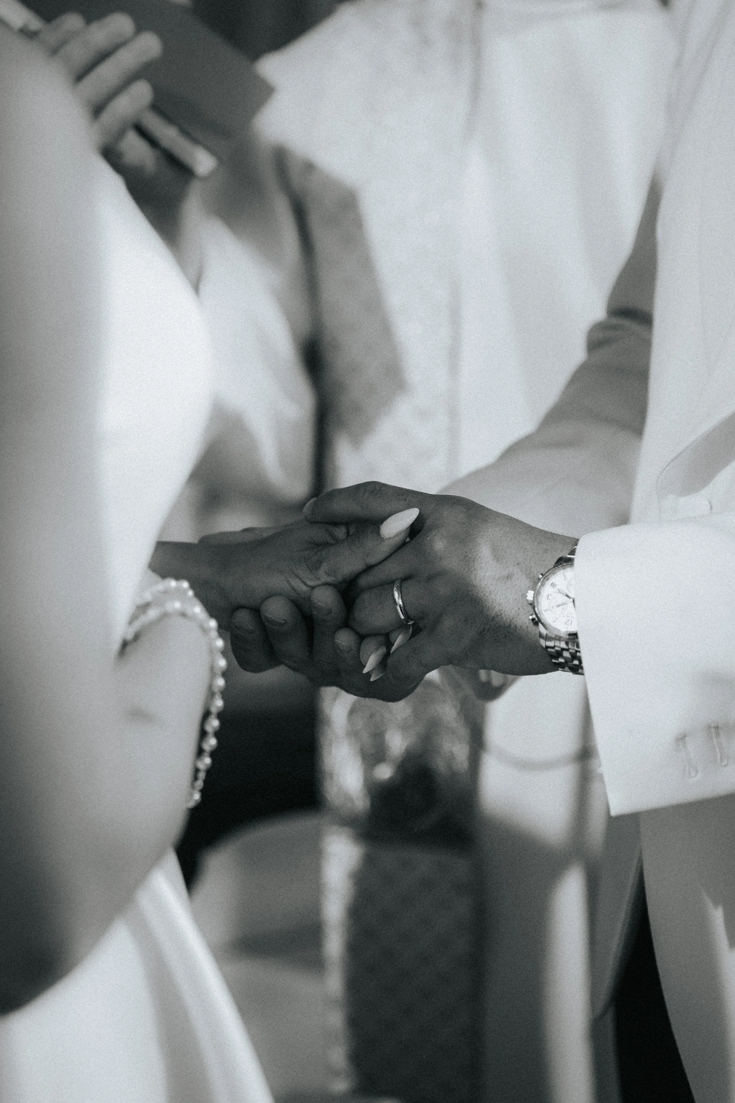 Close-up of two people holding hands, with one person wearing a watch and rings, and the other wearing a bracelet and rings, during a wedding or special ceremony.