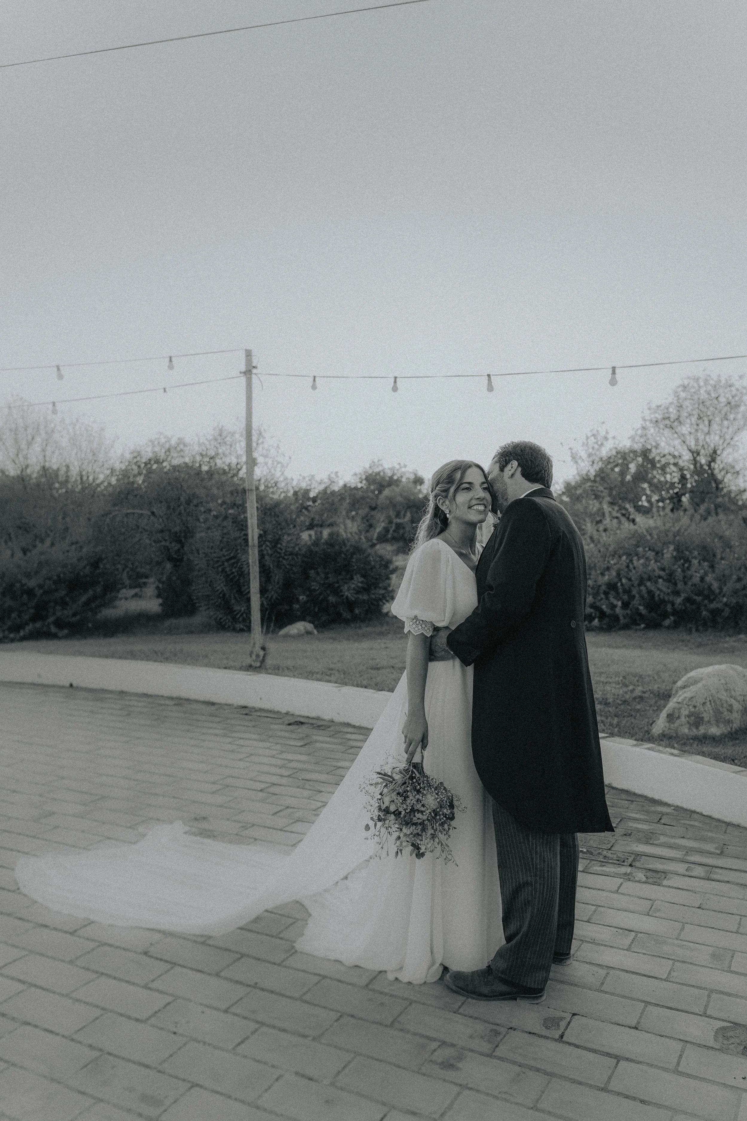 A black and white photo of a bride and groom standing close together outdoors, with the groom kissing the bride's temple. The bride is holding a bouquet of flowers and smiling. They are on a paved pathway, with trees, bushes, and a utility pole with 