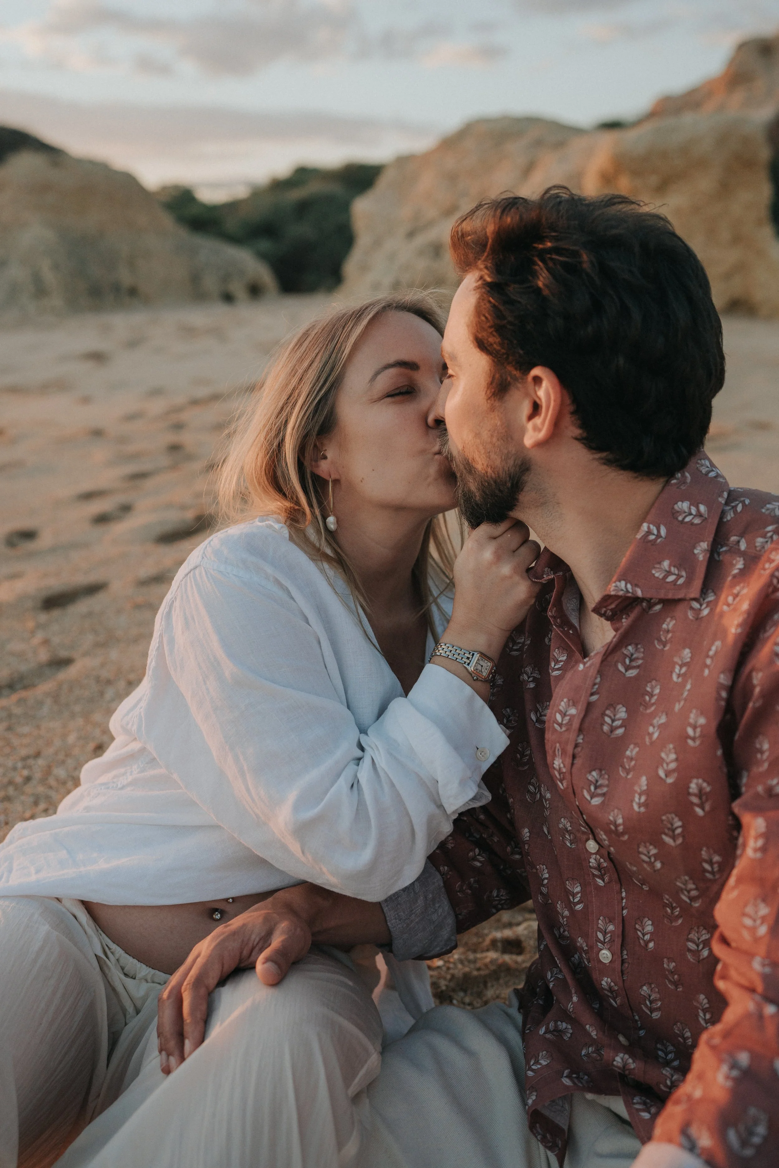 A couple kissing on a beach with large rocks in the background during sunset.