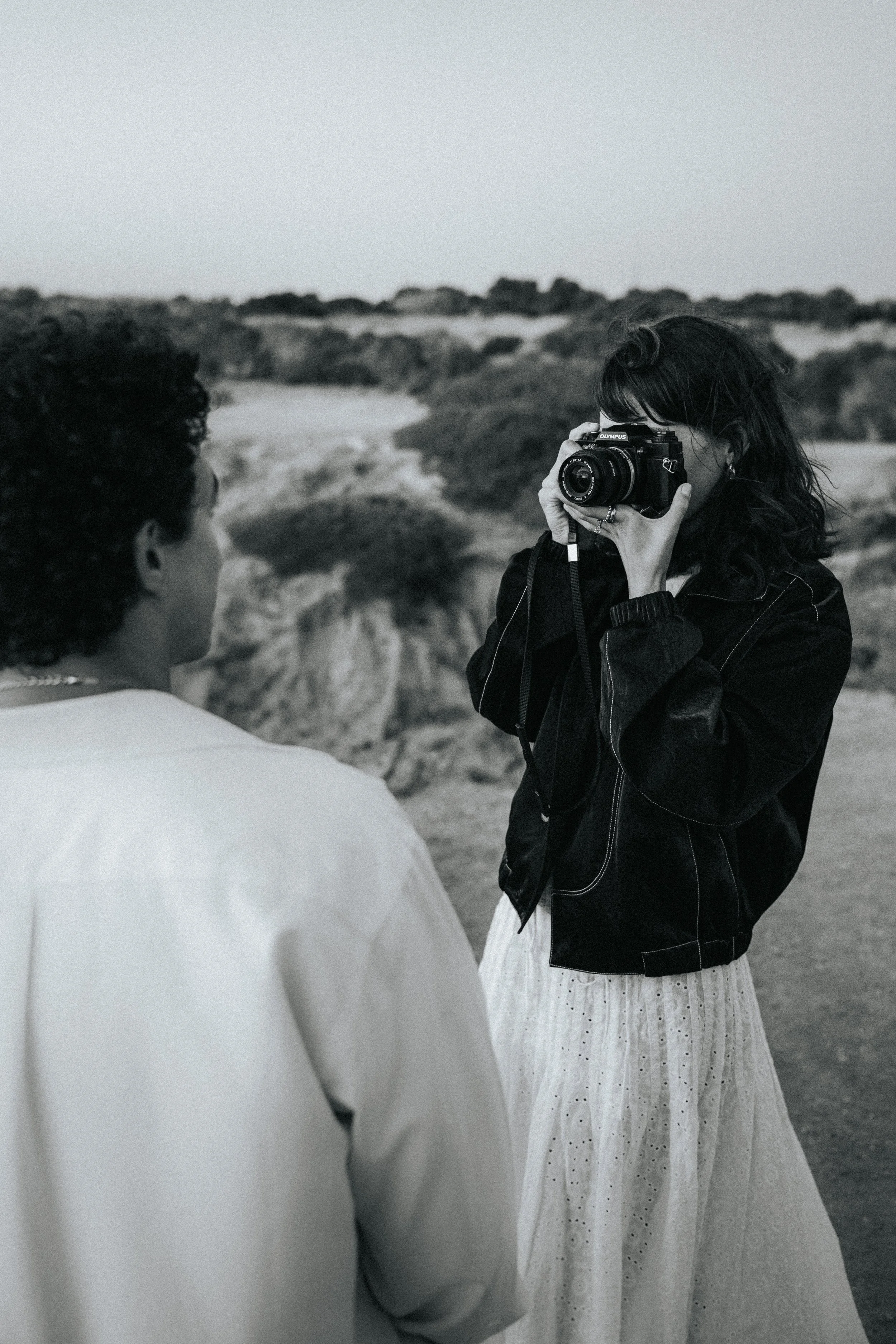 A woman with shoulder-length dark hair holding a camera to her face, taking a picture of a person in a white shirt, outside in a natural, possibly desert landscape with bushes and rocks.
