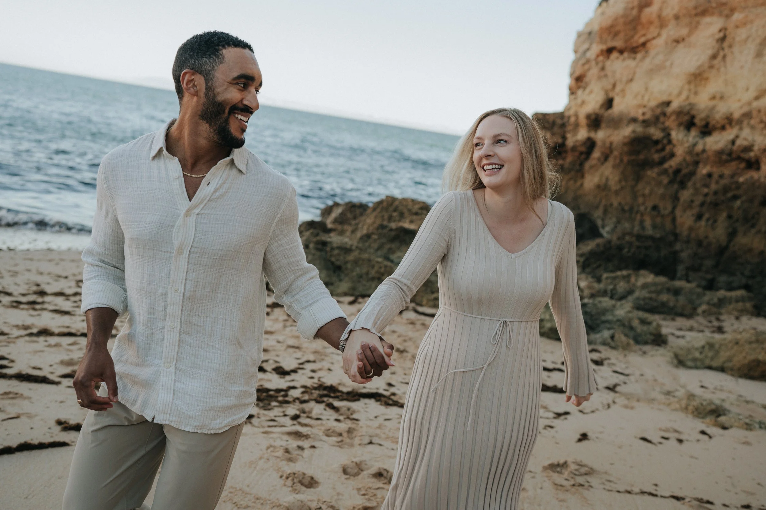 A couple holding hands and walking along the beach, smiling at each other with rocky cliffs and the ocean in the background.