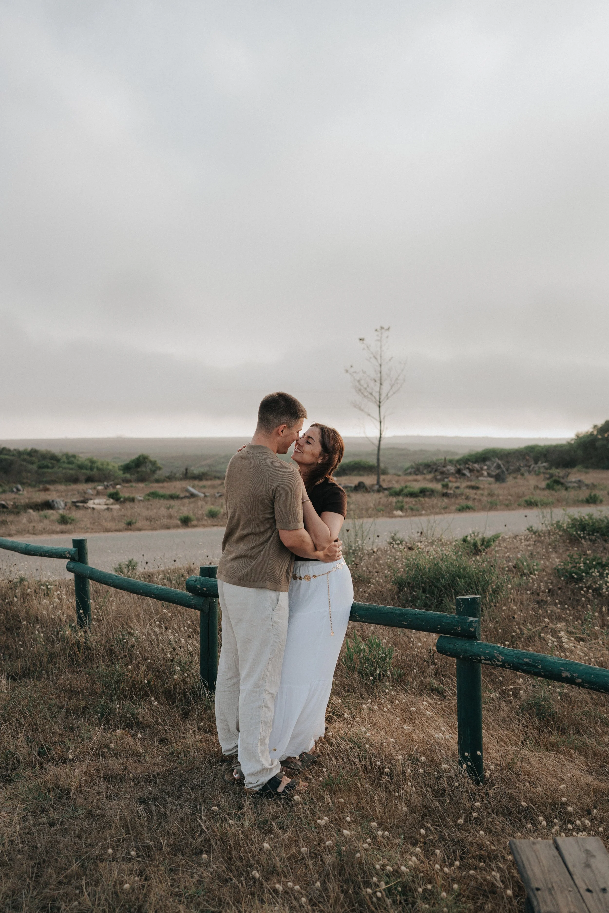 A couple stands close together by a green fence in an open field under a cloudy sky, with a single tree in the background.