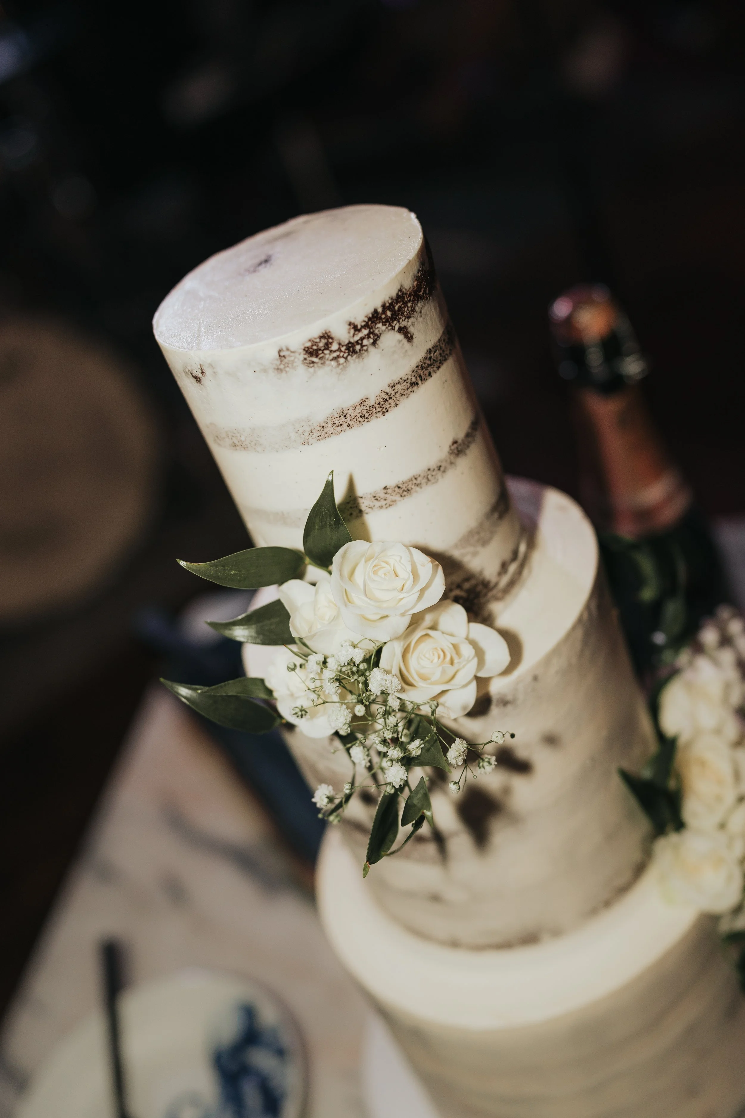 Close-up of a multi-tiered white wedding cake decorated with white roses and green leaves, with a blurred background.