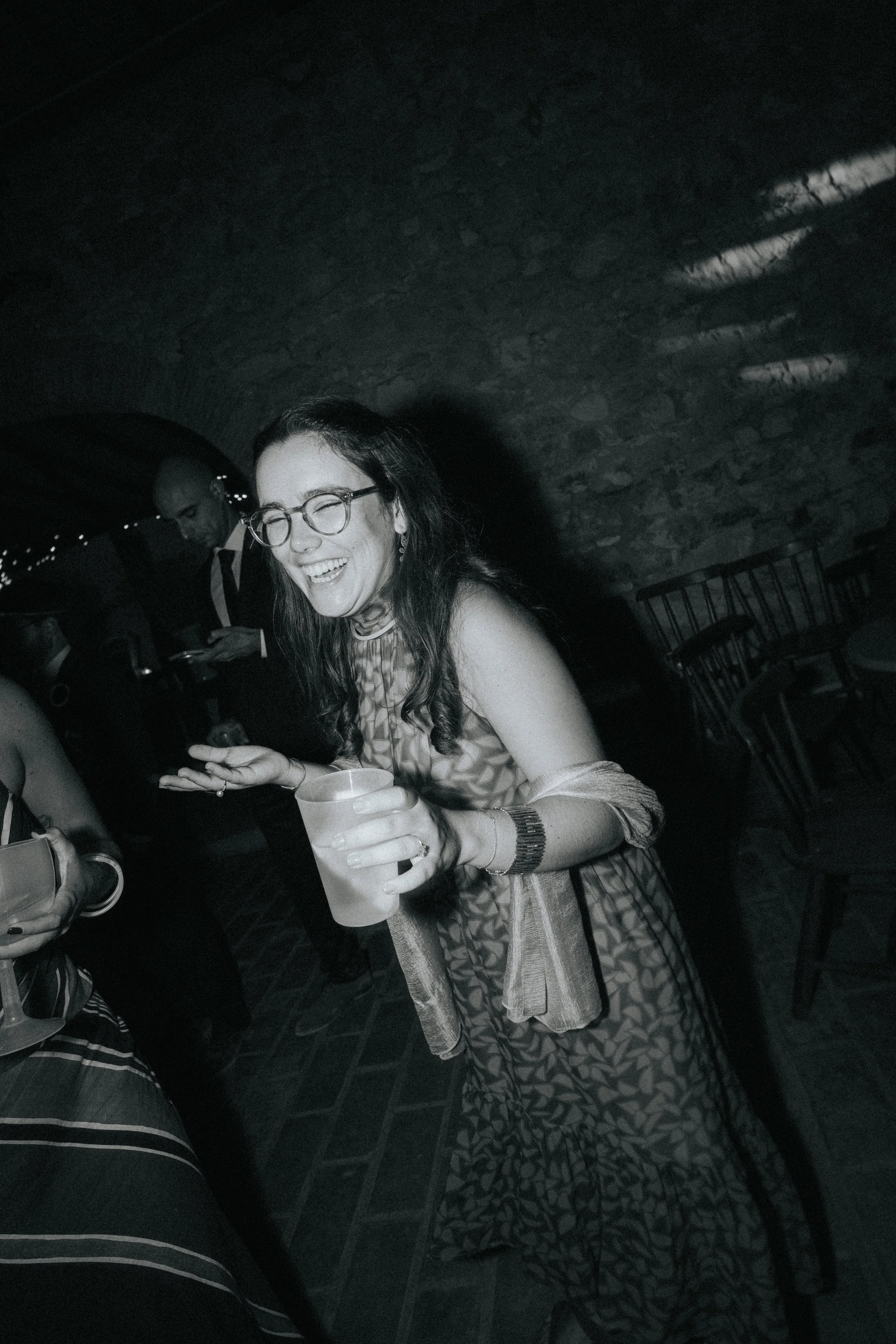 A woman wearing glasses and a patterned dress, smiling and holding a drink, in a dimly lit restaurant or bar setting.