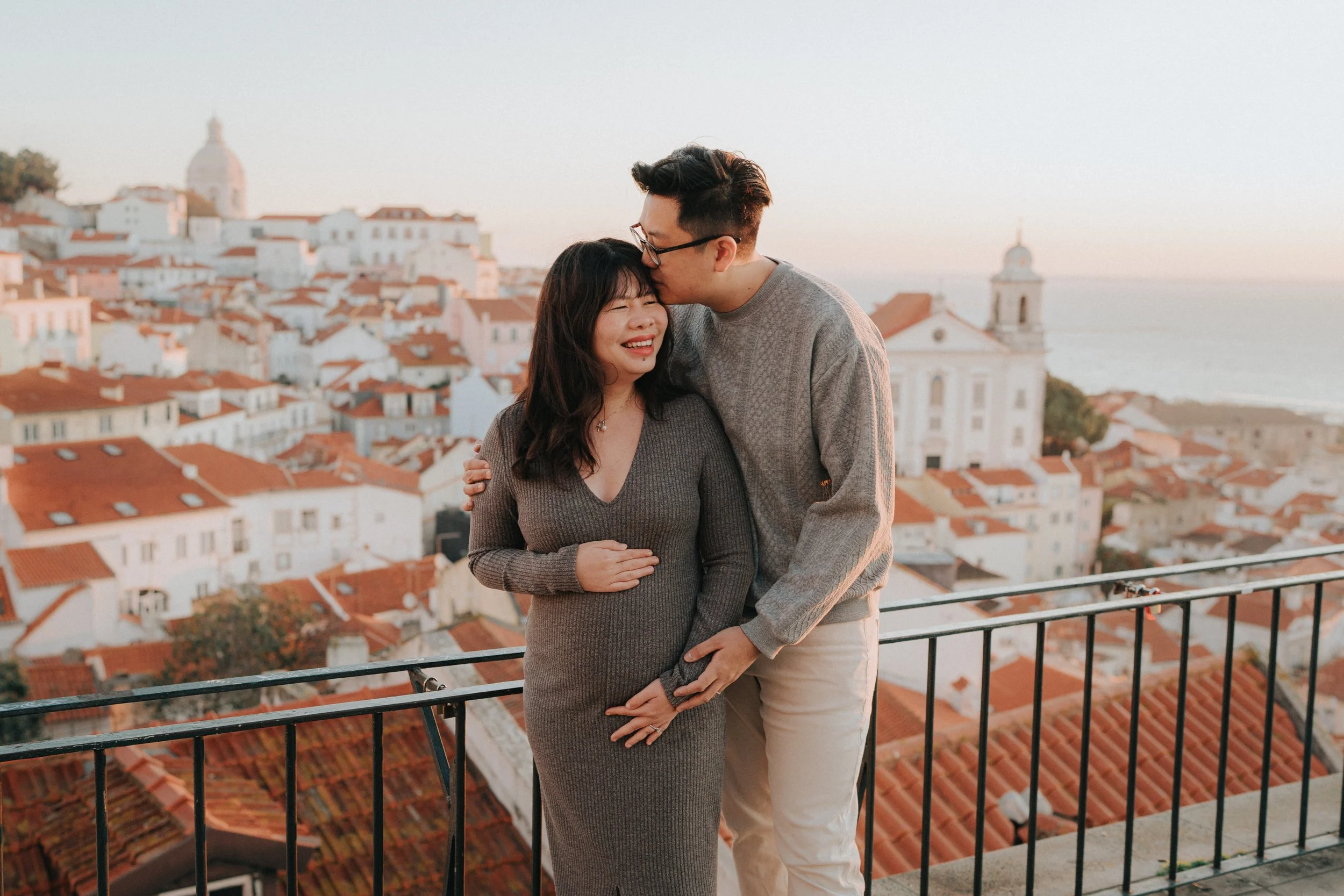 A couple embracing on a balcony overlooking a coastal town with red-tiled roofs and white buildings during sunset, the man is kissing the woman on her forehead.