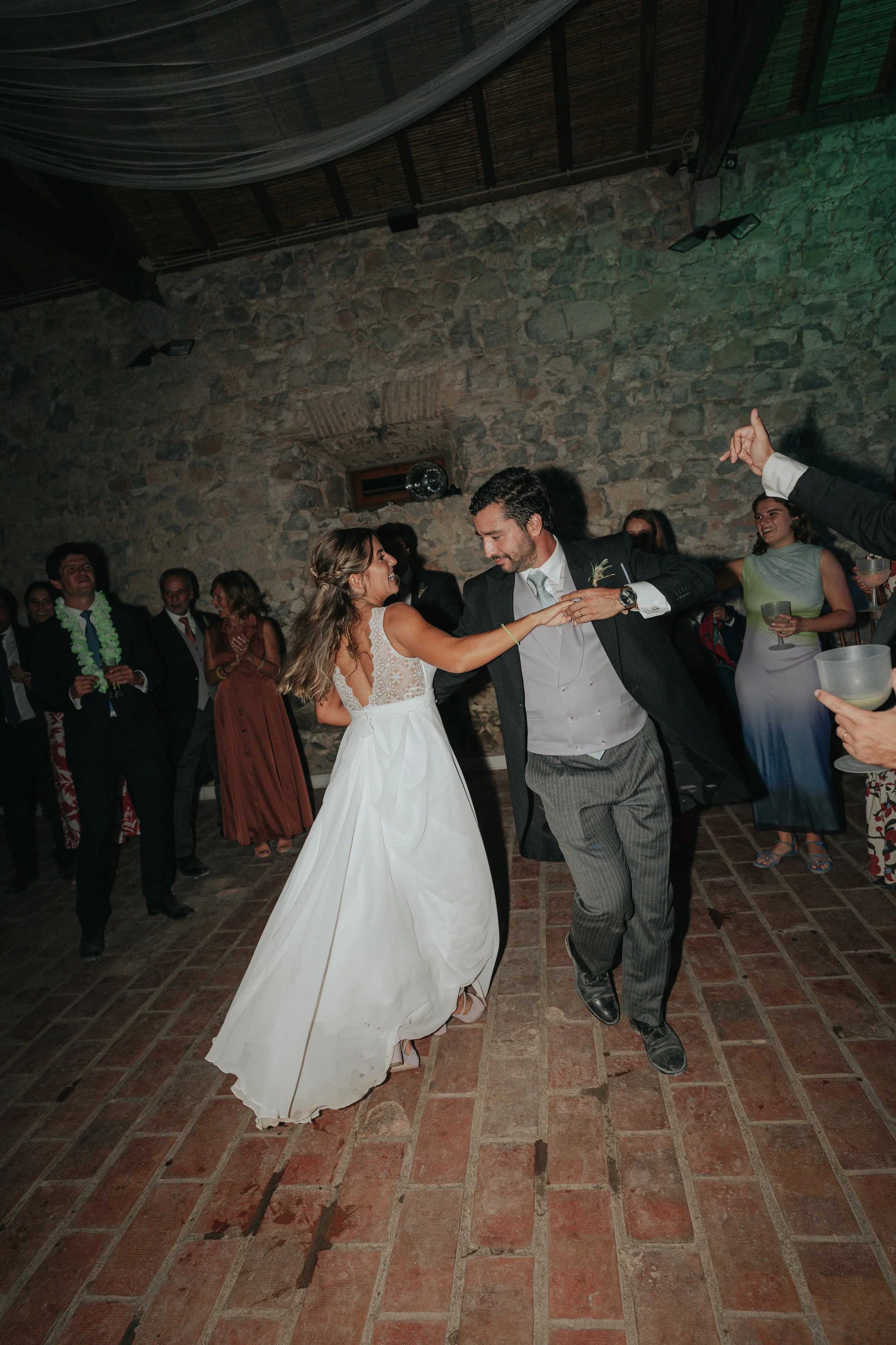 Couple dancing at a wedding reception with guests watching and celebrating in the background, in a rustic venue with stone walls and brick flooring.
