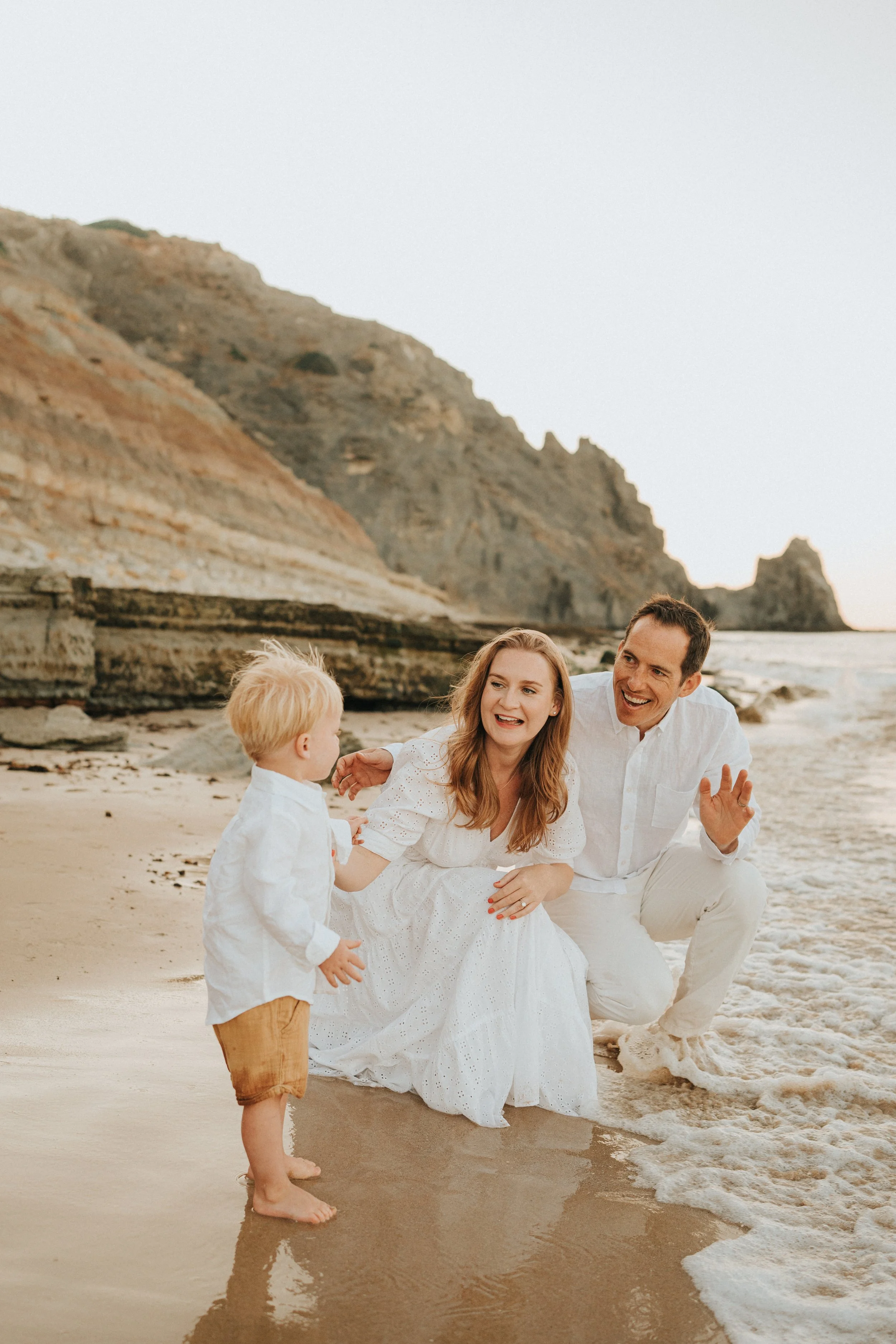 A family of four, dressed in white, enjoying time together on a sandy beach near rocky cliffs at sunset. The mother, father, and two children are smiling and playing by the water.