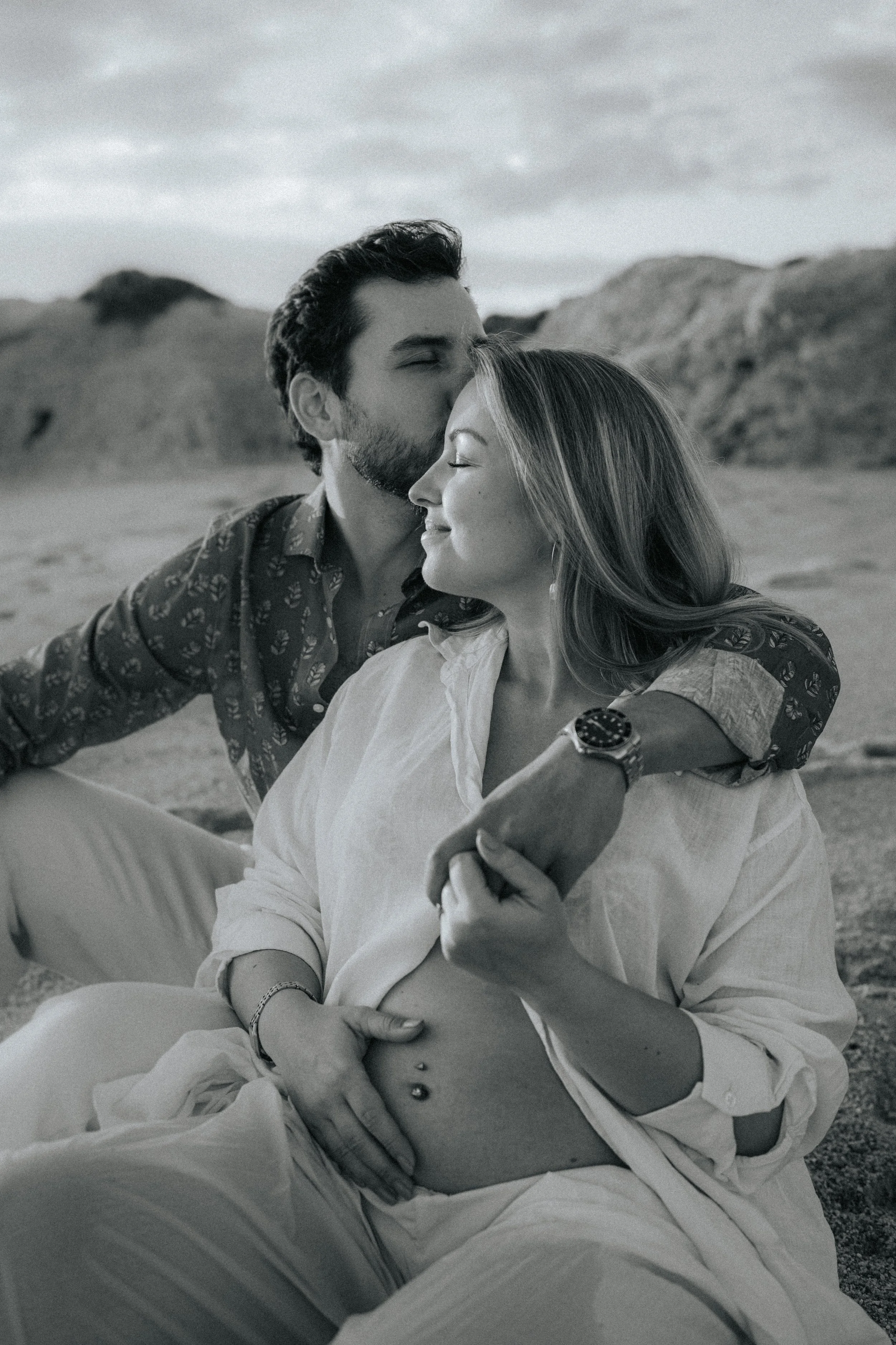 A black and white photo of a young couple sitting outdoors, embracing and smiling with their eyes closed. The man is kissing the woman's forehead, and they appear happy and peaceful. The background shows a mountainous landscape under cloudy skies.