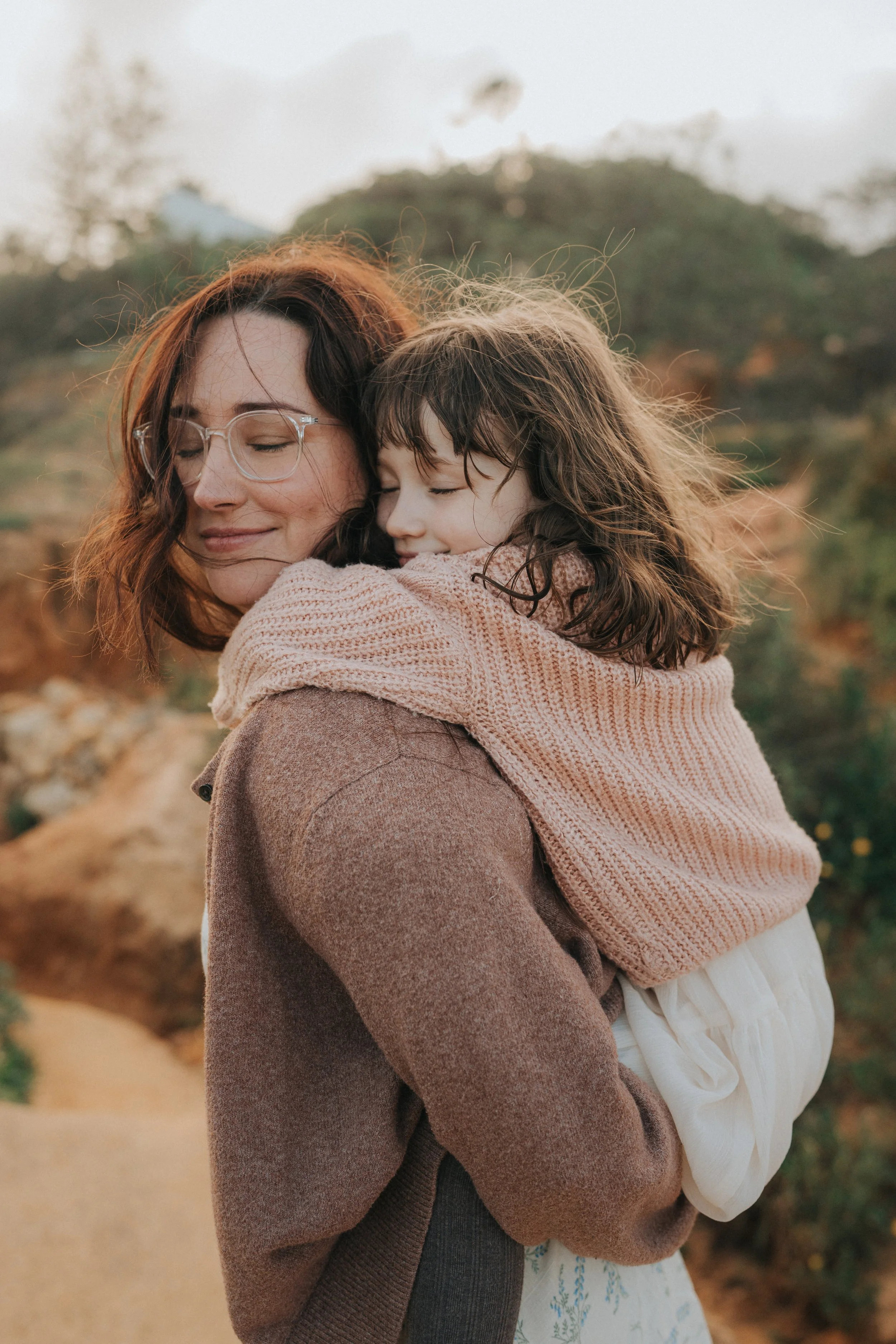 A woman with red hair and glasses hugging a young girl with dark hair and a knitted pink sweater, outdoors in a natural setting with blurred trees and a path in the background.