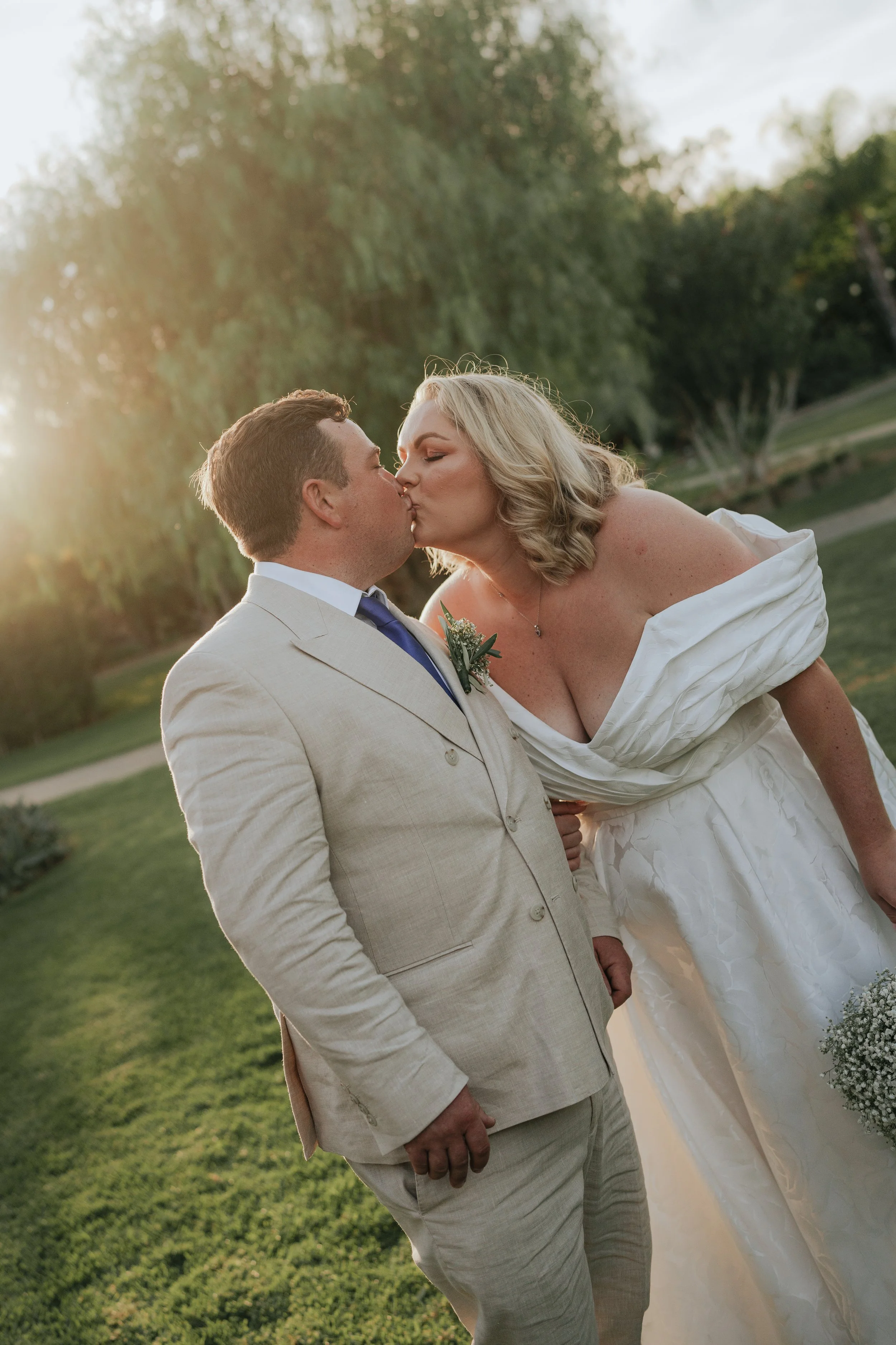 A bride and groom share a kiss outdoors during sunset, with the bride in a white wedding gown and the groom in a light-colored suit, surrounded by trees and grass.