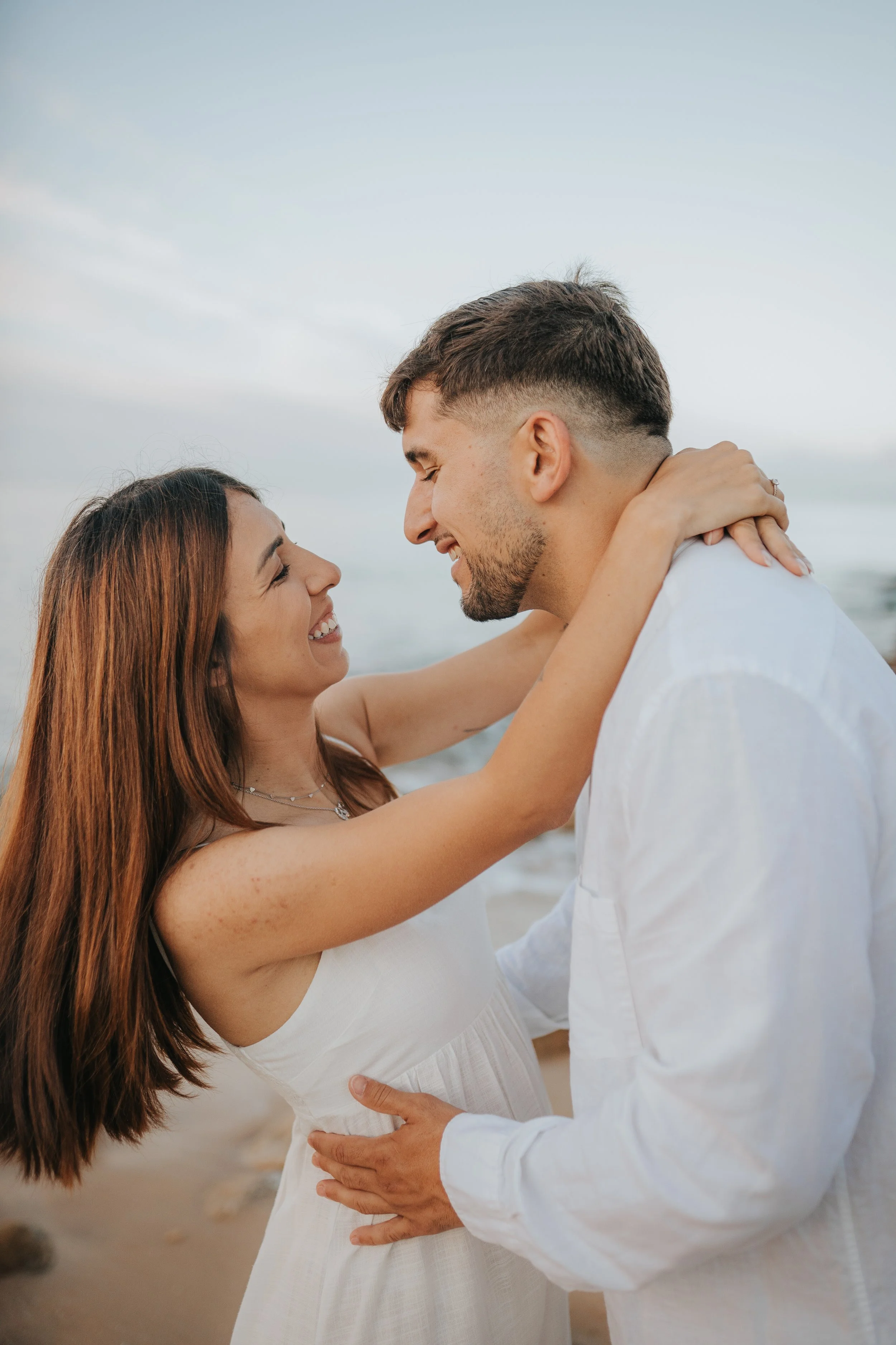 A happy couple at the beach, holding each other close and smiling, with the ocean in the background.