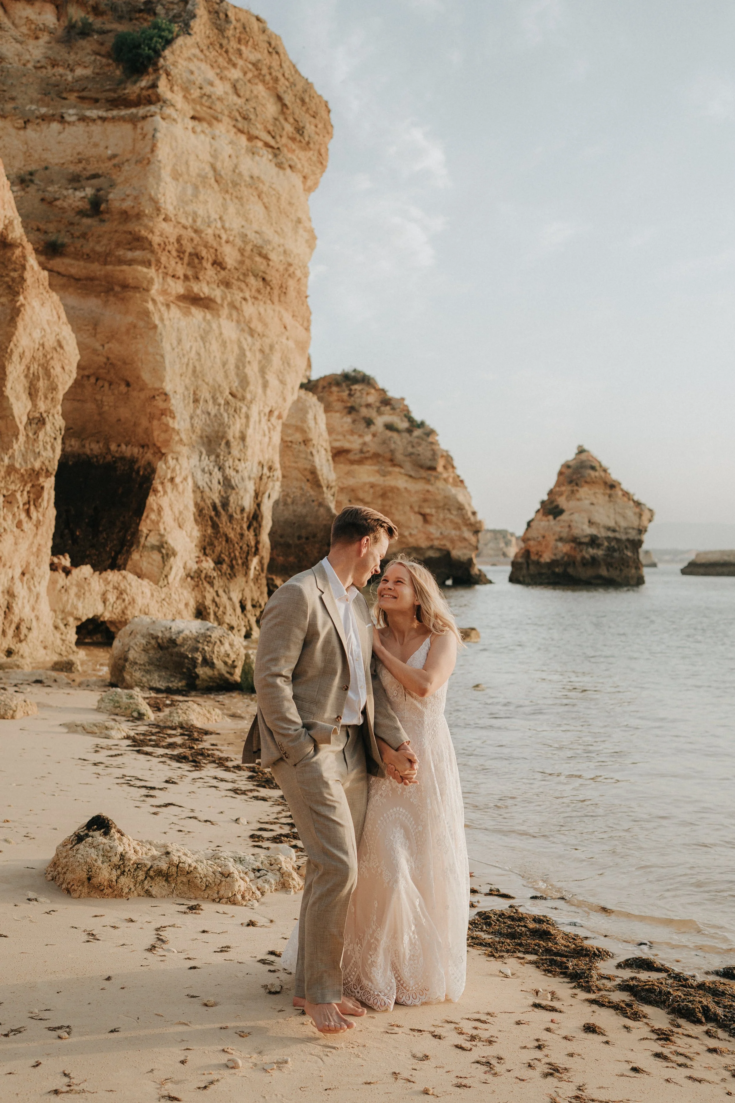 A couple holding hands on the beach near large rock formations, with the woman in a white wedding dress and the man in a light brown suit, smiling at each other.
