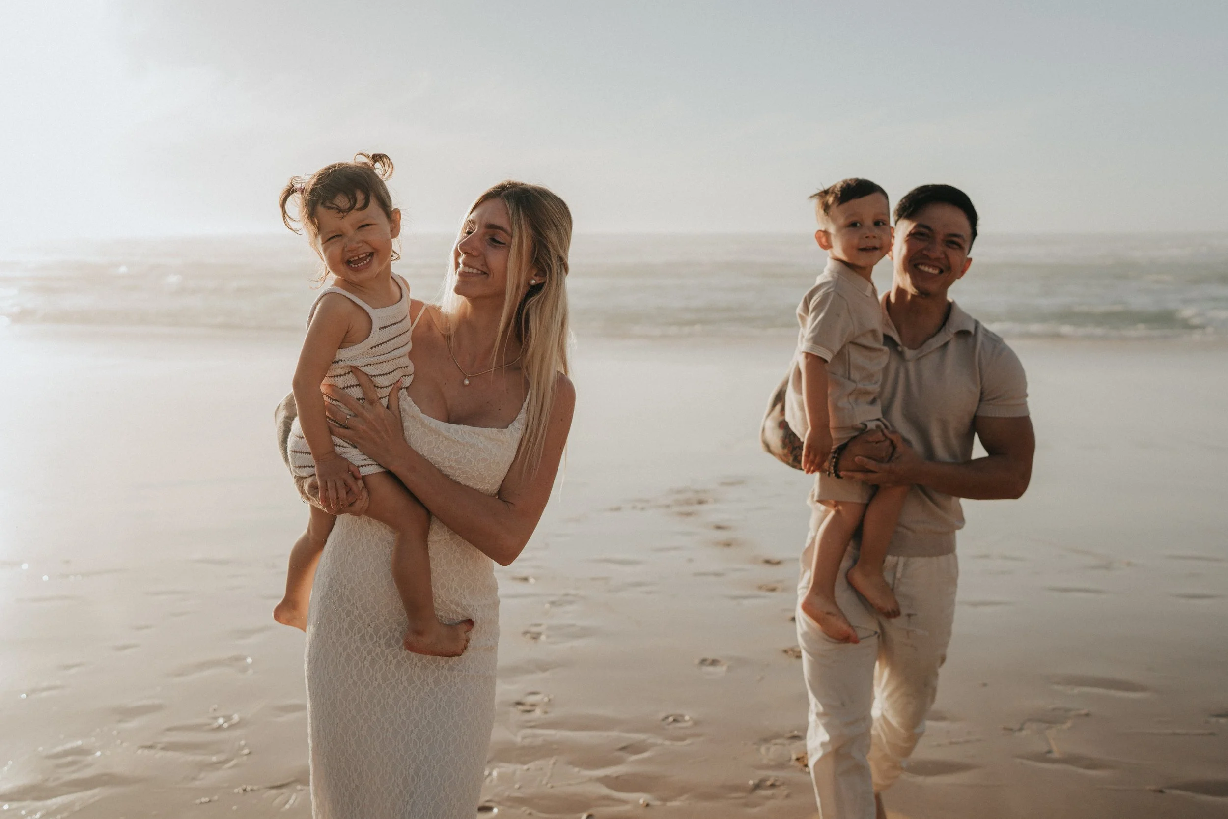 A family of four enjoying a day at the beach, with two parents and two young children, all smiling and walking along the shoreline.