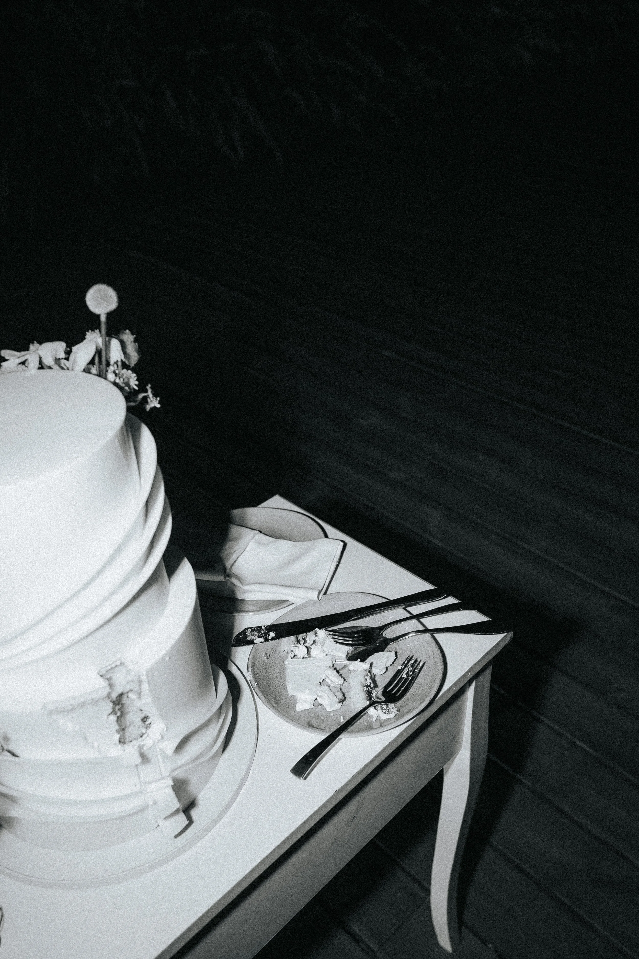 A partially eaten white cake with multiple layers on a white table, with a cake knife nearby. There are empty plates, some with leftover cake pieces and utensils, on the table. The background has a dark wood or panel wall.