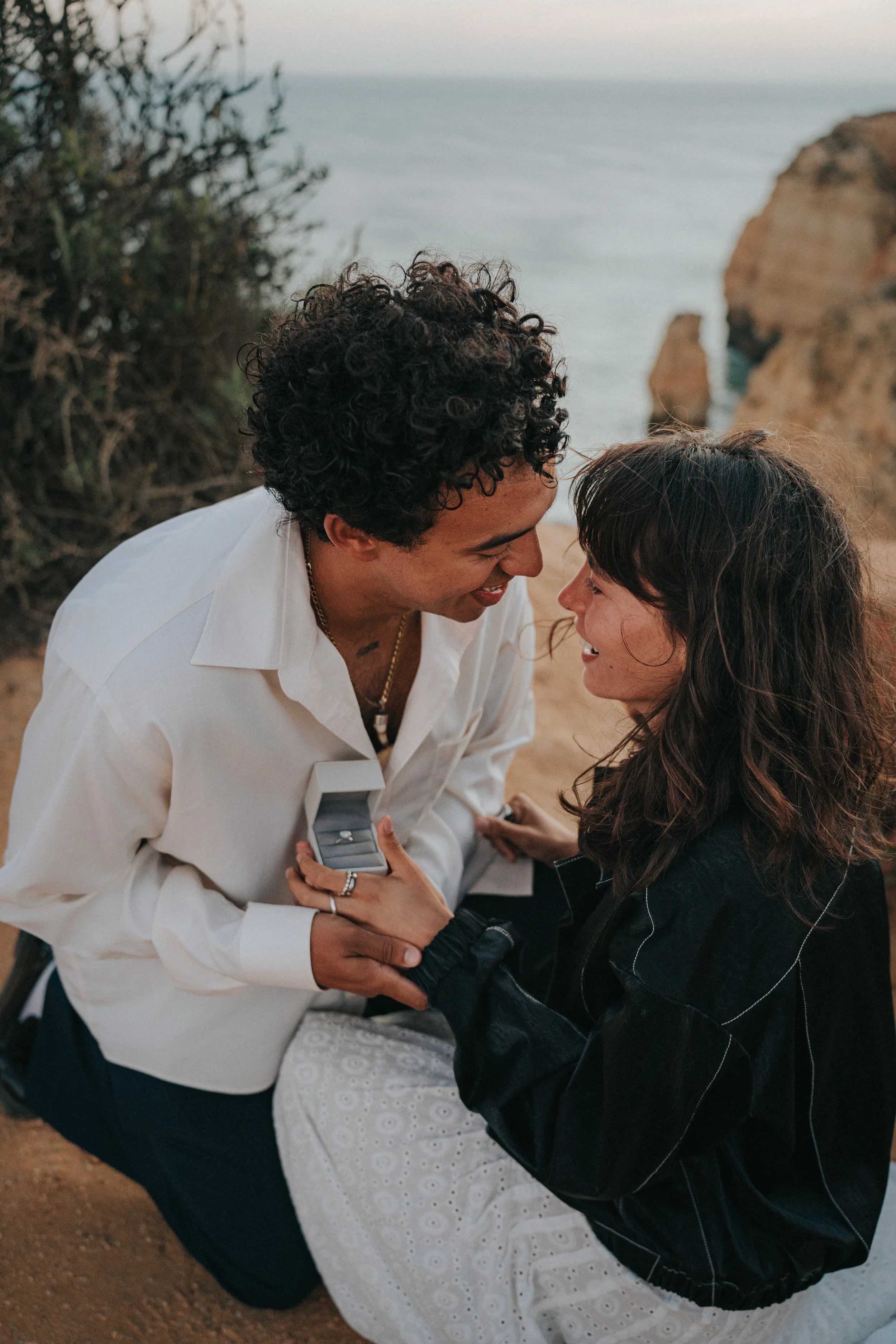 A man proposing to a woman outdoors near the ocean during sunset, with rocky formations in the background.