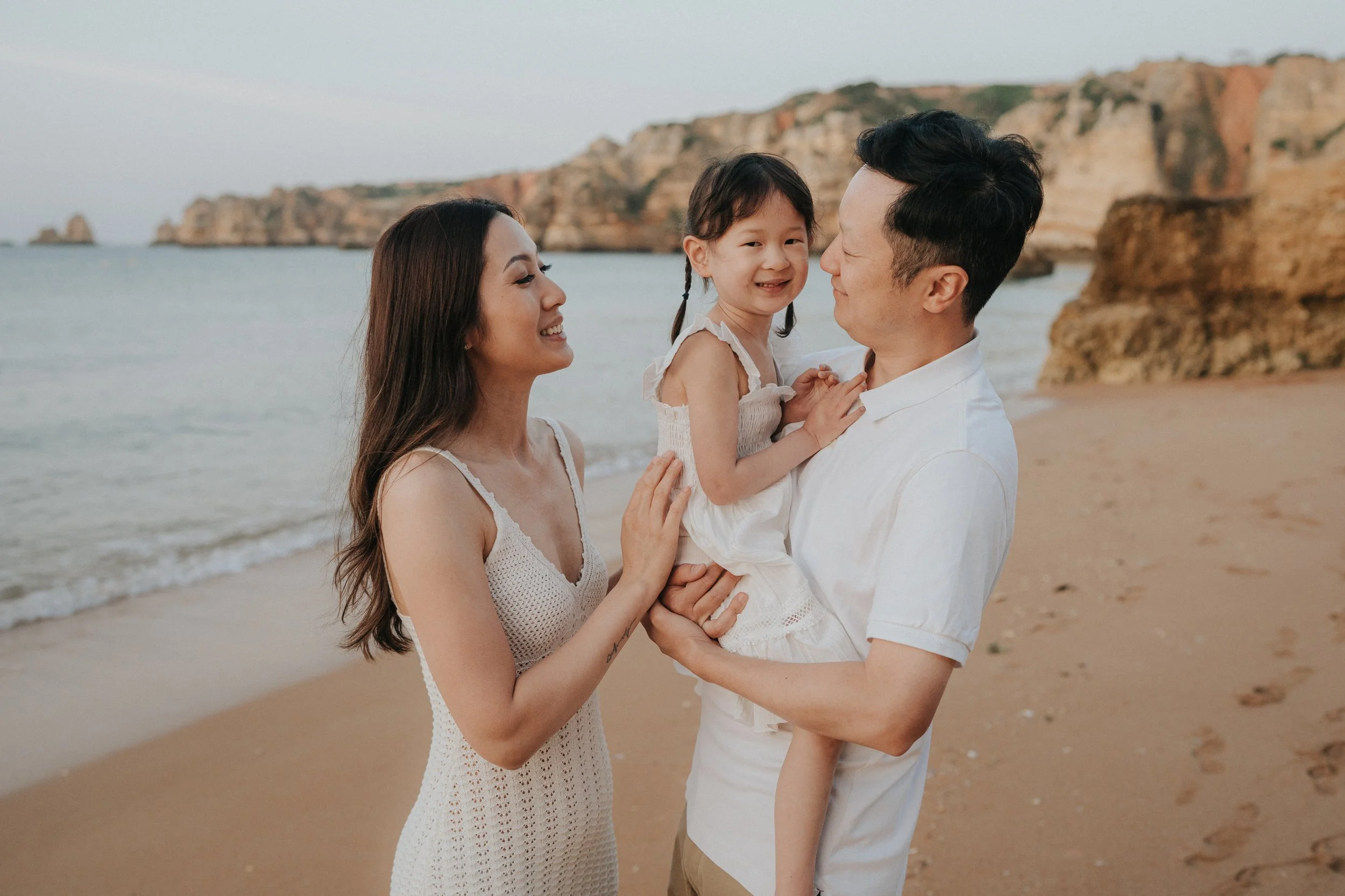 A family of three at the beach, with rocky cliffs in the background. The woman, man, and young girl are smiling and holding each other, enjoying a sunny day.