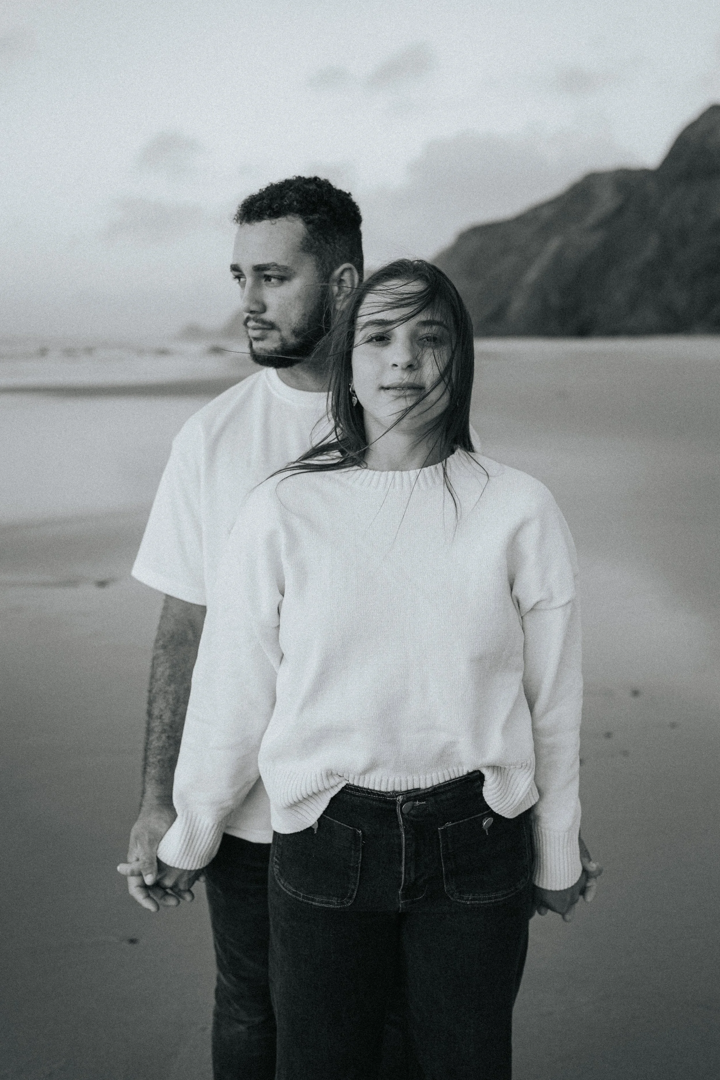 Black and white photo of a man and woman holding hands on a beach, with a large mountain in the background. The woman is in front with her hair blowing in the wind, and the man is slightly behind her, looking to the side.