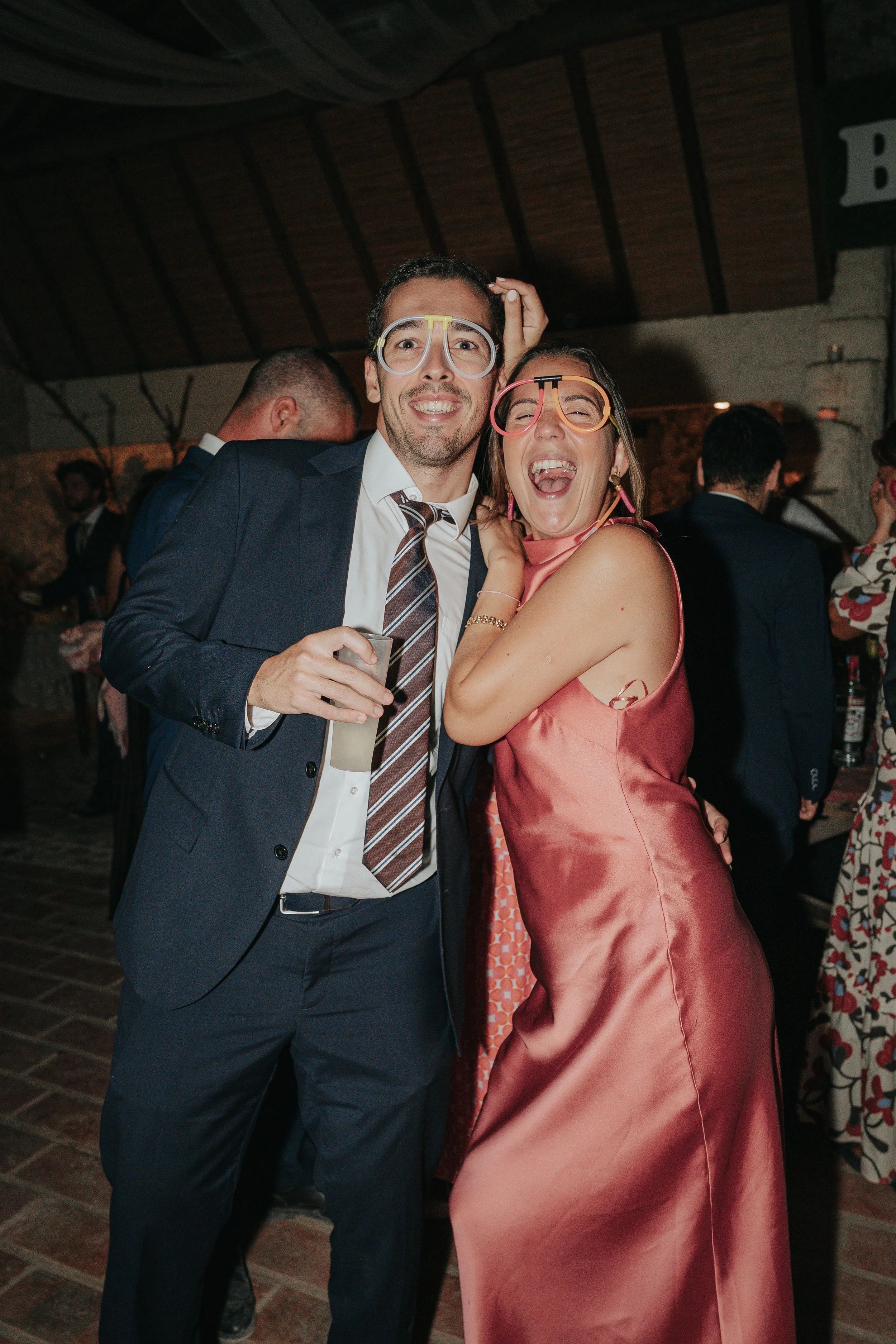 A joyful man in a suit and a woman in a pink dress wearing colorful glasses celebrating at a party with other people in the background.