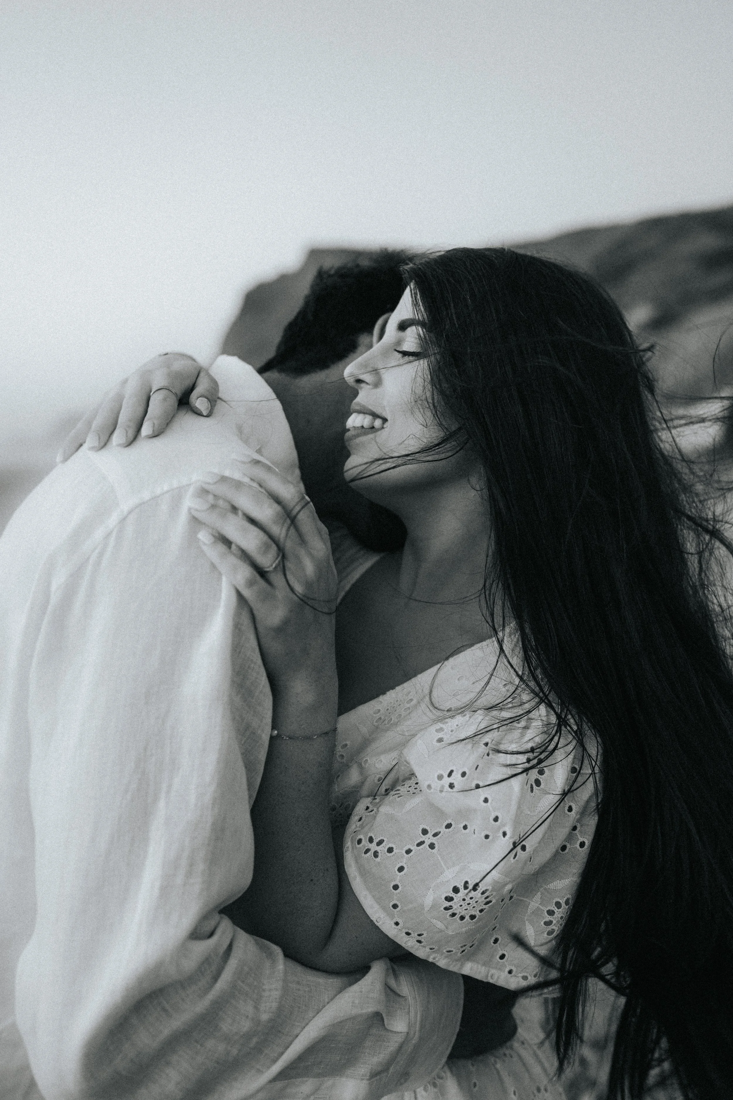 A black and white photo of a woman smiling with closed eyes, hugging a man from the side, both seemingly enjoying a tender moment.