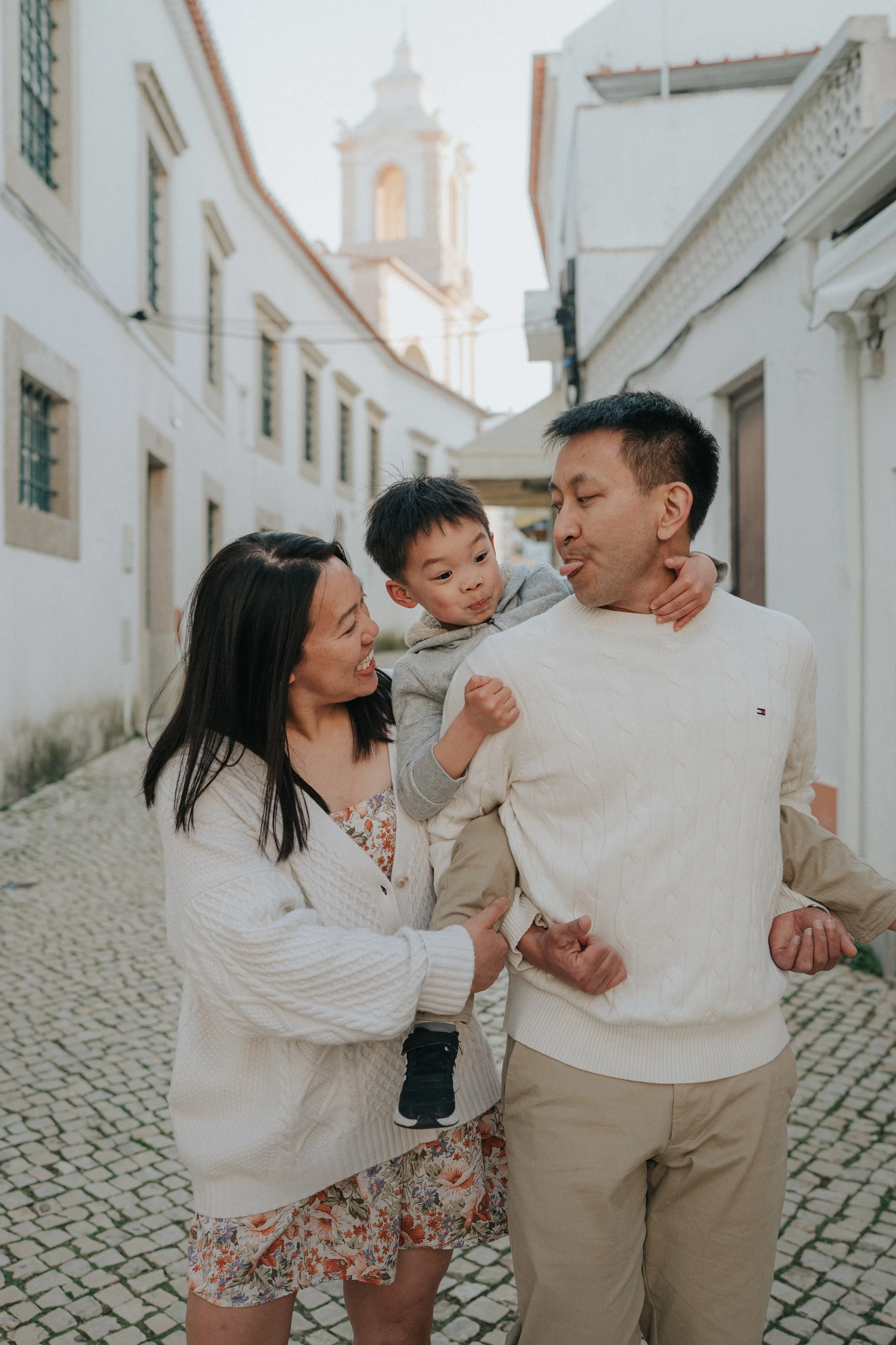 A family of four enjoying a walk on a cobblestone street in a European city, with historic white buildings and a church tower in the background.