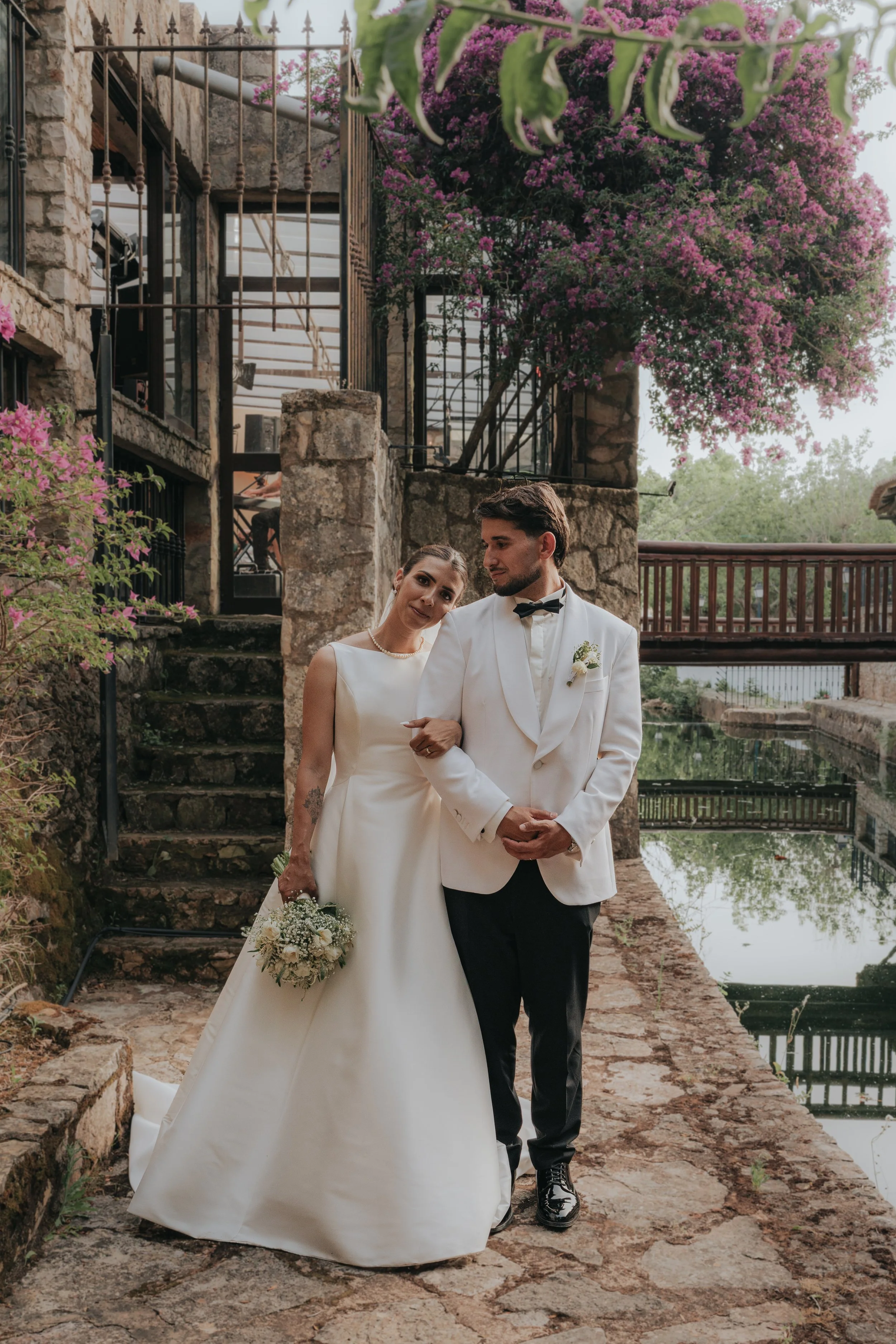A bride and groom standing outdoors on a stone path, with a stone and iron fence, blooming pink flowers, and a bridge over water in the background.