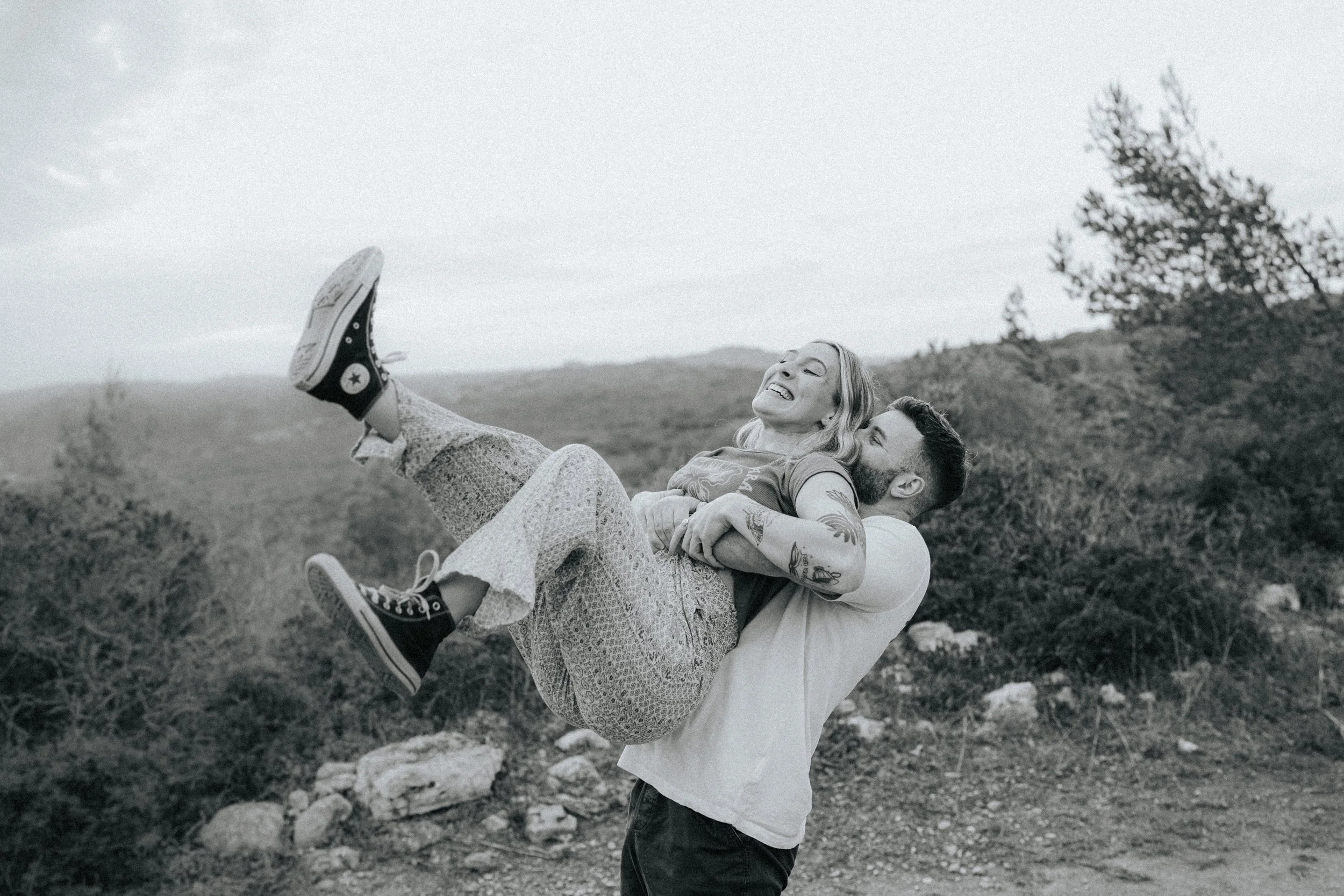 A man lifting a woman in the air outdoors in a grassy field, both smiling and appearing joyful, with hills and trees in the background.