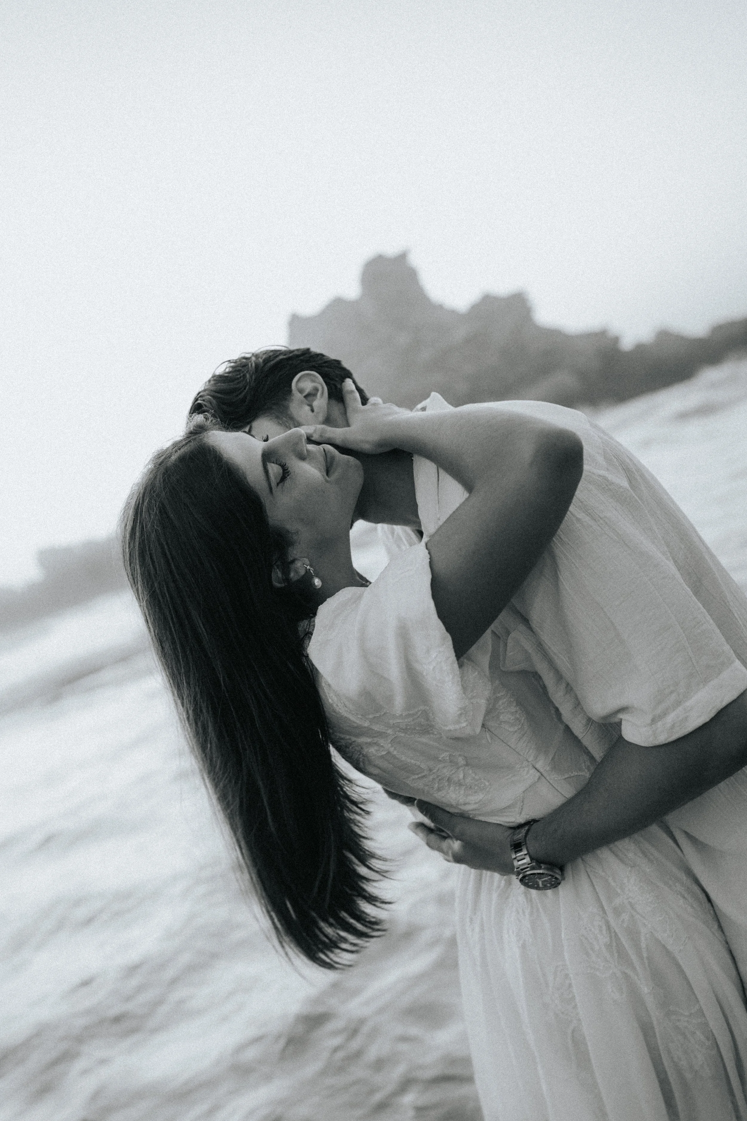 A black-and-white photo of a couple dancing closely on the beach, with rocky formations in the background.