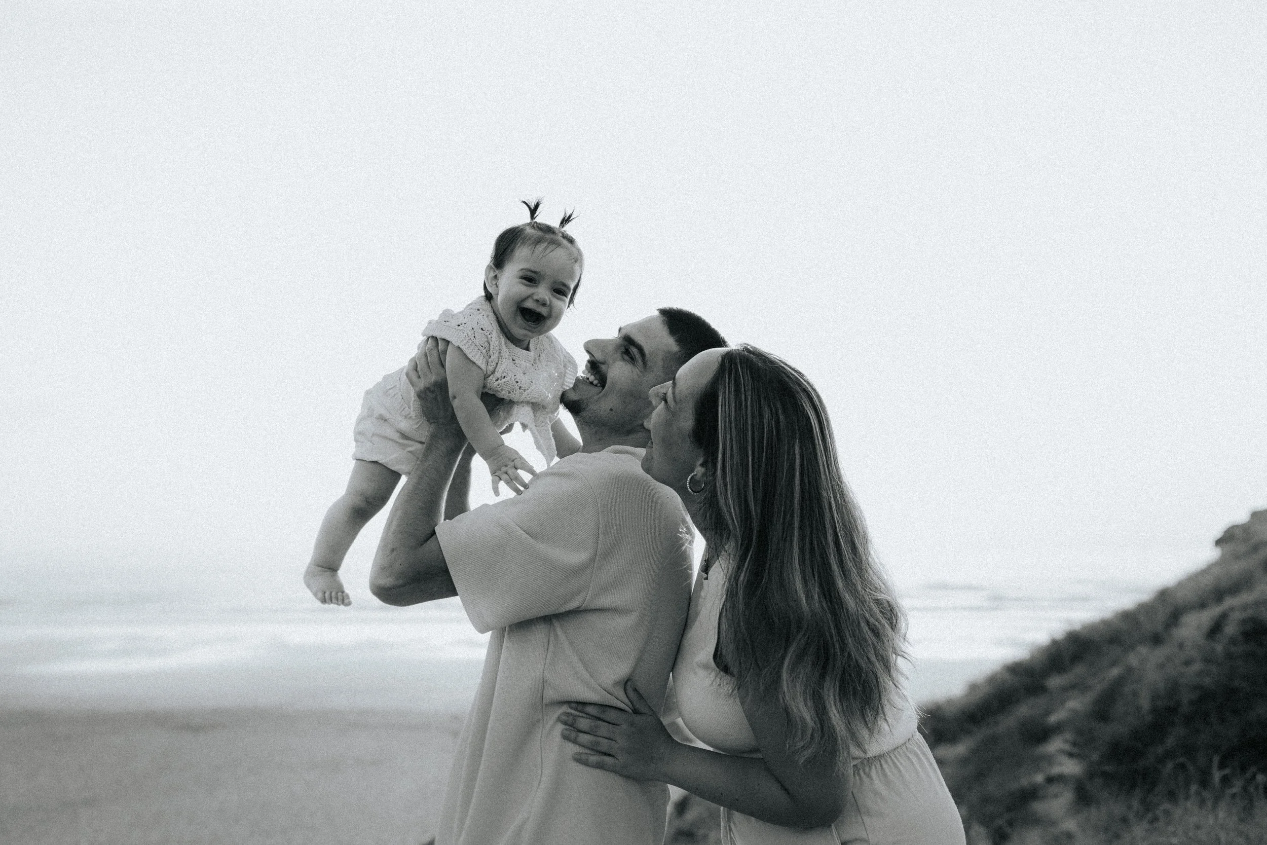 A woman lifting a smiling toddler girl outdoors near a beach or coastal area.