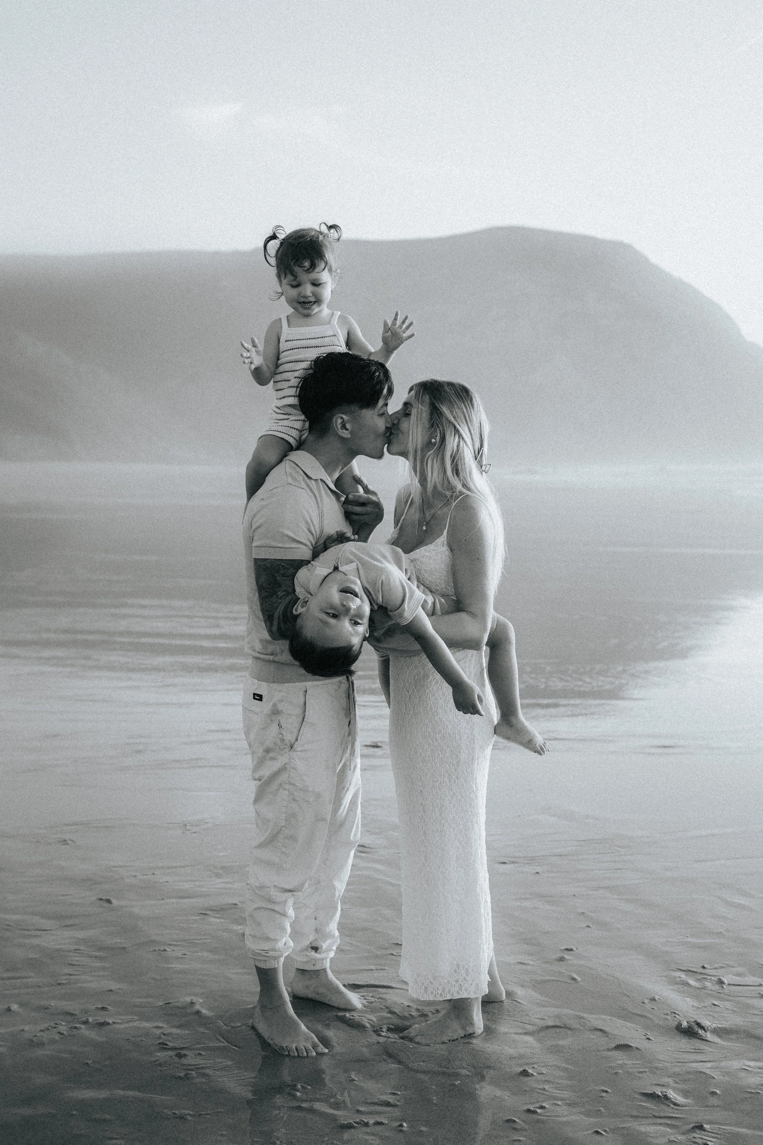 A black and white photo of a family on the beach. A man is holding a woman, and they are kissing. A young girl is sitting on the man's shoulders, smiling and waving, while a boy is upside down with his head hanging near the woman's dress, also smilin