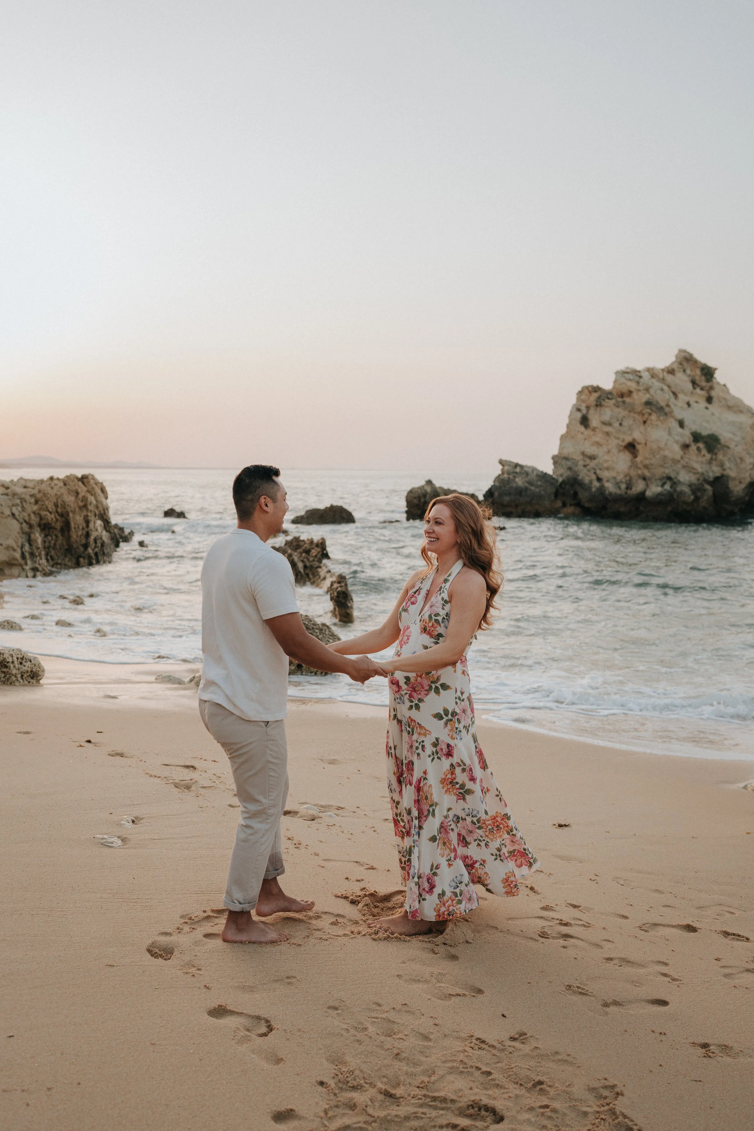 A couple holding hands and smiling at each other on a sandy beach during sunset, with rocky formations and the ocean in the background.