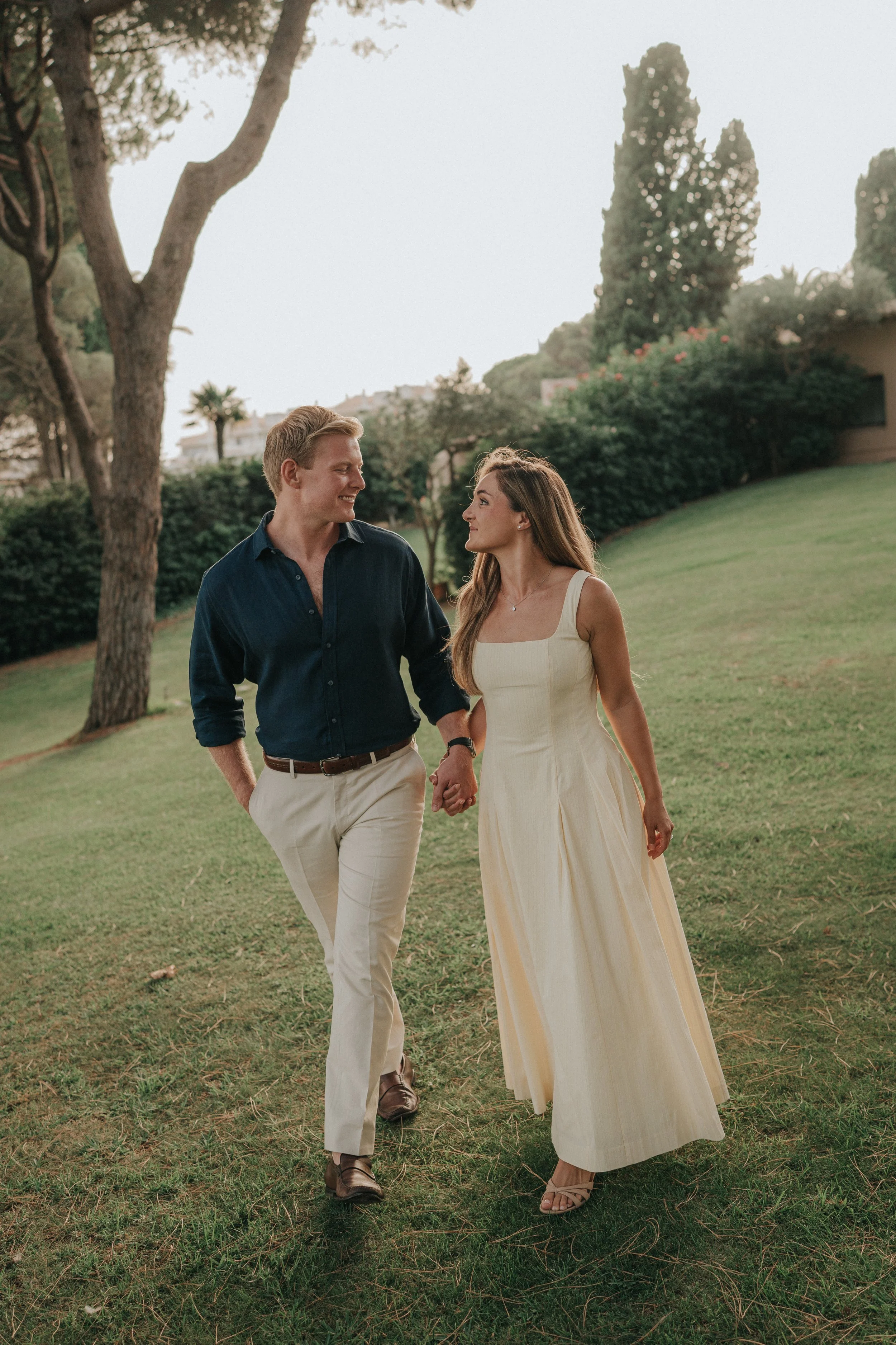 A young couple walking hand-in-hand outdoors in a park or garden during the daytime, surrounded by trees and greenery, with a woman in a long, light-colored dress and a man in a dark blue shirt and light-colored pants.