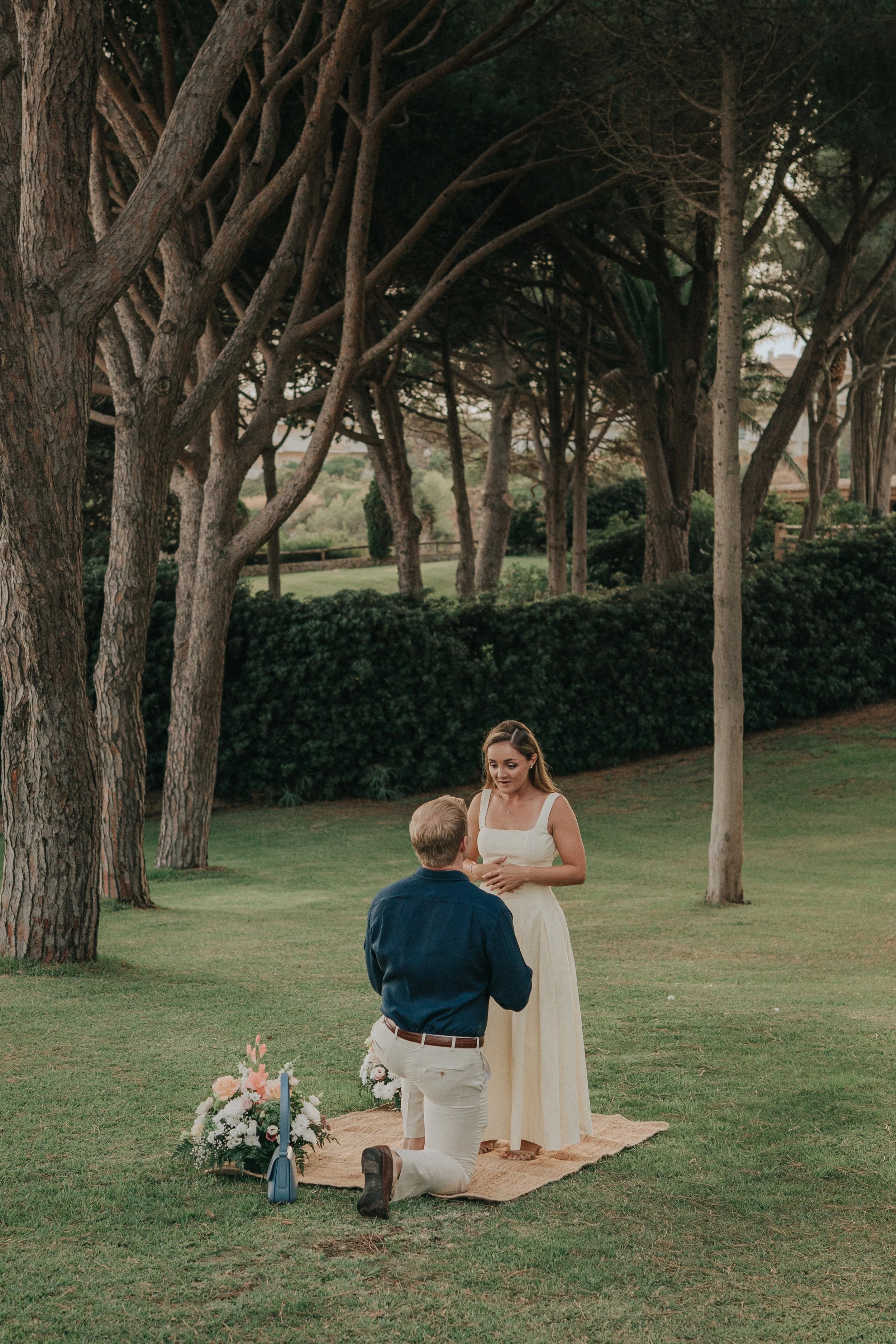 A couple getting married outdoors in a park, kneeling on a rug with flowers and candles, with tall trees and green bushes in the background.