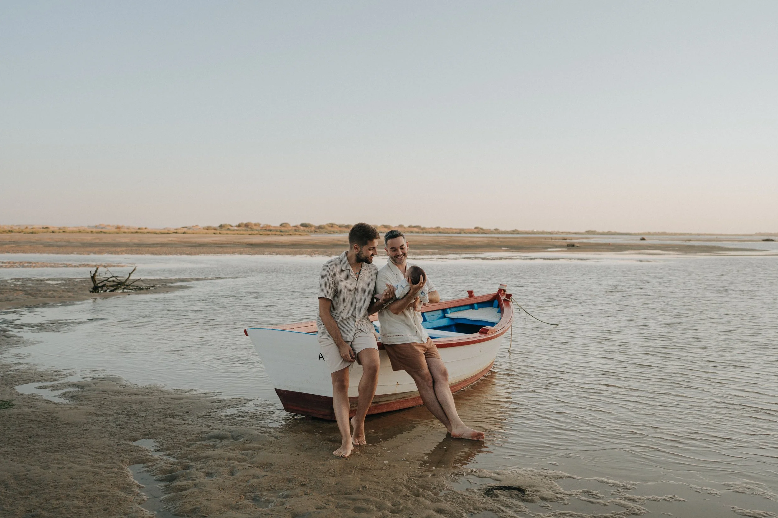 Two men sitting on the edge of a small boat at the water's edge, holding a newborn baby, with a calm river and distant shoreline under a soft sky.