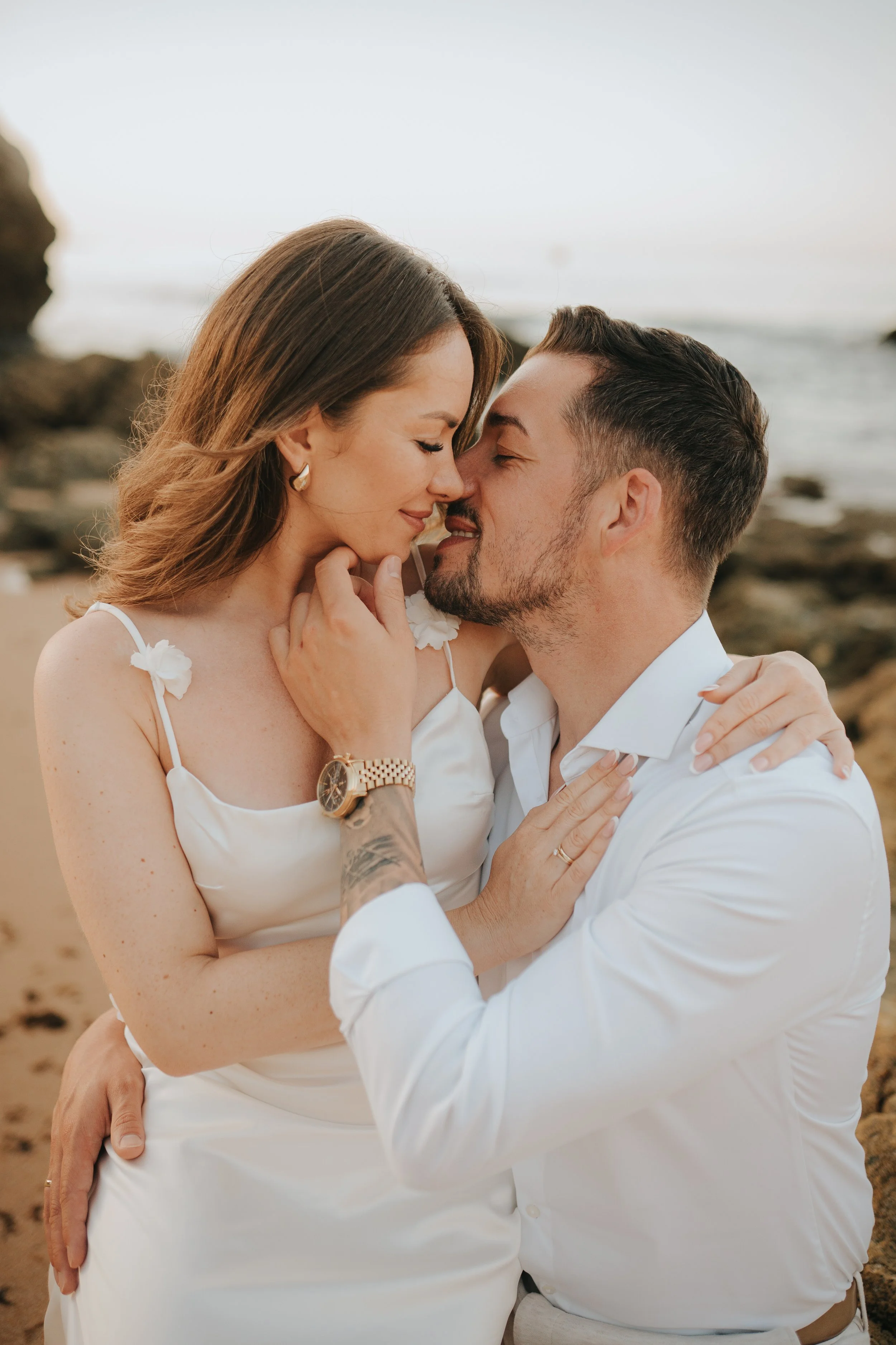 A couple embracing and touching foreheads on the beach, both with eyes closed, wearing white clothing.