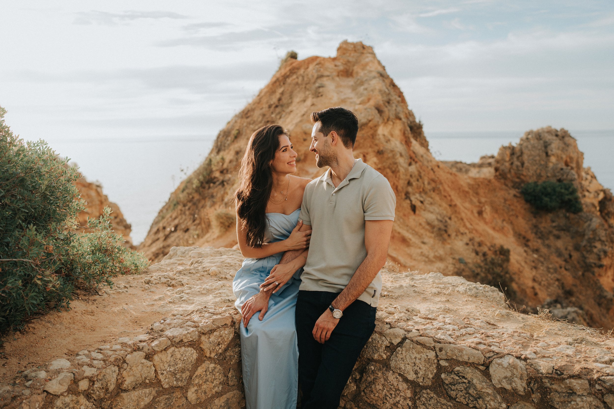 A smiling couple sitting on a rocky ledge in front of orange cliffs, with the sea and cloudy sky in the background.